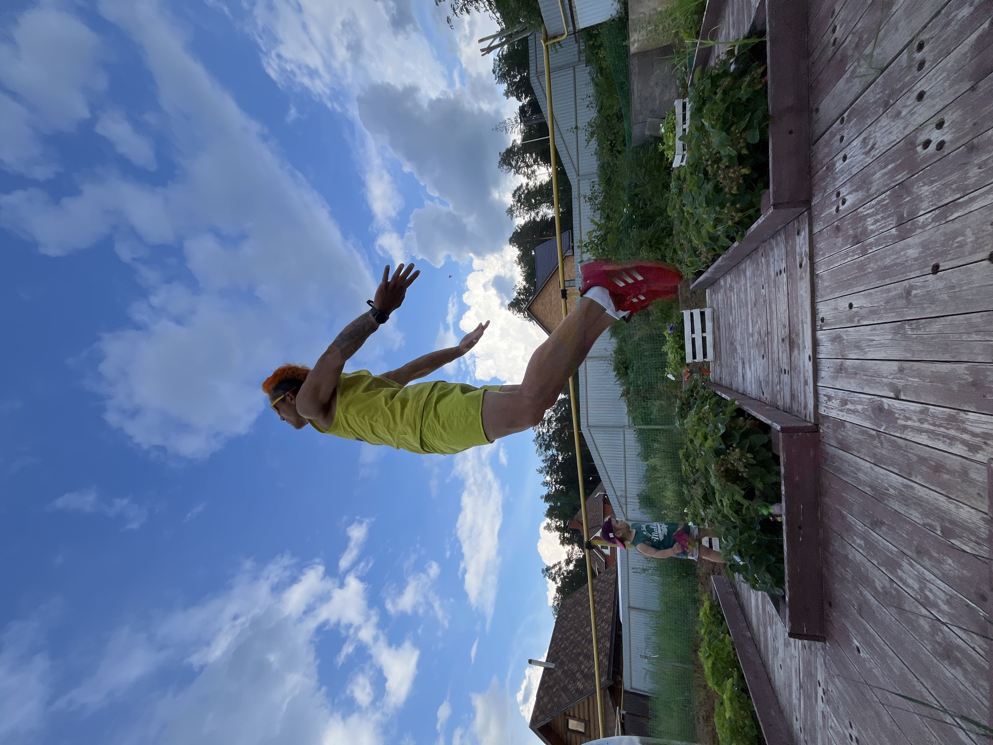 In Chaikovsky, Russia, an individual with bright orange hair, wearing a yellow tank top and shorts, is captured mid-air in a dynamic jump. They are in a low-angle shot, with outstretched arms and legs, appearing to be leaping towards the sky. The background features a cloudy blue sky with scattered white clouds. Below the jumping figure, a child in a green tank top and a purple baseball cap stands near raised garden beds filled with green plants. The scene is set in an outdoor environment, possibly a backyard or garden, with wooden decking in the foreground and a fence and trees visible in the distance. Buildings, appearing to be houses, are also present in the background.