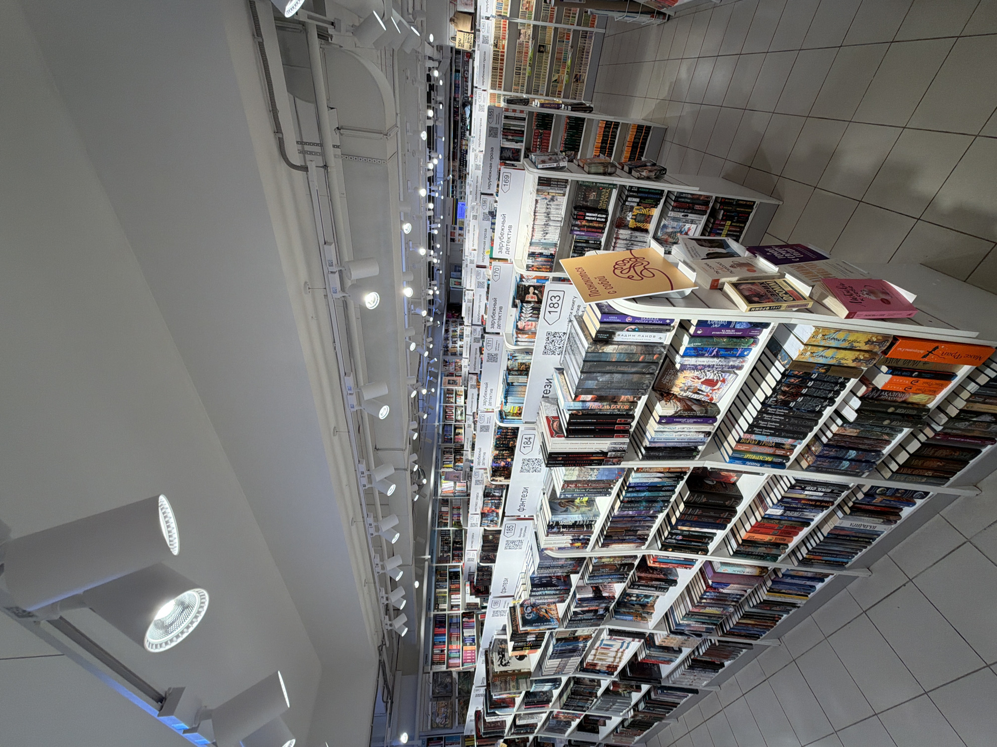 This indoor shot depicts a bookstore aisle in Izhevsk, Russia. The focus is on the towering shelves filled with books. The books are neatly stacked in rows, sorted by genre, indicated by category labels. The books' spines are visible, creating a visually appealing pattern of colors and titles. The shelving units are white, contrasting with the colorful book covers. Above, a ceiling with numerous lighting fixtures illuminates the scene. The floor is tiled, adding to the clean and organized appearance. The overall impression is one of a well-stocked and carefully curated bookstore.