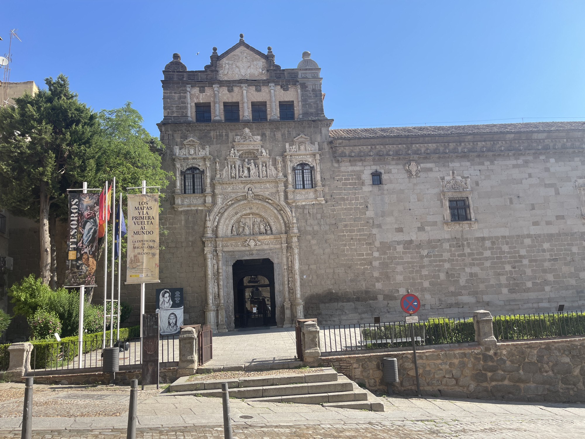 In Toledo, Spain, the image captures the entrance to the Church of Santo Tomé under a clear blue sky. The church features a grand stone facade with intricate carvings around its main entrance, which is a dark, imposing archway. Above the entrance, a decorative stone frieze depicts figures and architectural elements. The building's walls are constructed of large, rough-hewn stones.
To the left of the entrance, several banners are displayed on flagpoles. One banner, partially visible, has the word "TESOROS" at the top and a depiction of a religious scene. Another banner, in Spanish, reads "LOS MAPAS Y LA PRIMERA VUELTA AL MUNDO" (The Maps and the First Circumnavigation of the World) and mentions "LA EXPEDICIÓN DE MAGALLANES-EL CANO" (The Expedition of Magellan-El Cano). Below these banners, two framed pictures are mounted on a stand; one shows two women, and the other features a depiction of Christ.
In front of the church, a stone wall with a black metal railing encloses a green, manicured area with bushes. Steps lead up to the church entrance from a cobblestone walkway. A no-parking sign with a red circle and blue arrow is visible to the right of the entrance, near a trash bin and stone retaining wall. The foreground shows a cobblestone street with bollards and a sidewalk. The overall scene suggests a sunny day in a historic city.