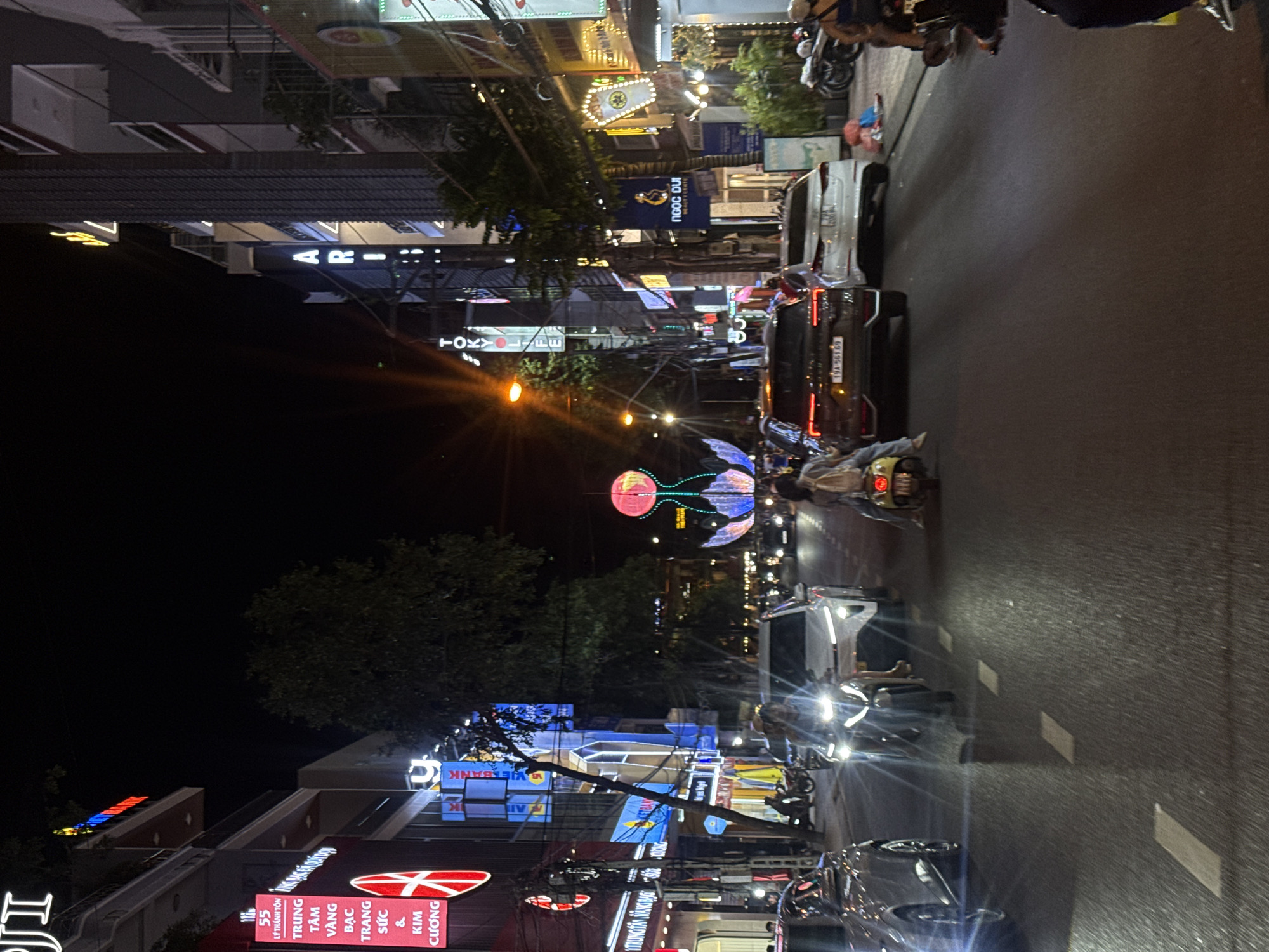 The image depicts a street scene at night in Nha Trang Ward, Vietnam. The street is illuminated by various neon signs and streetlights, casting a vibrant glow on the surroundings. Several vehicles, including cars and motorbikes, are on the road, with some motorbikes occupied by individuals. Buildings line both sides of the street, displaying a mix of commercial establishments with illuminated signage. The architecture suggests a busy urban environment. Decorative lights in the shape of a lotus flower are visible in the distance, adding to the festive atmosphere. The overall scene captures the energetic nightlife of a Vietnamese city.