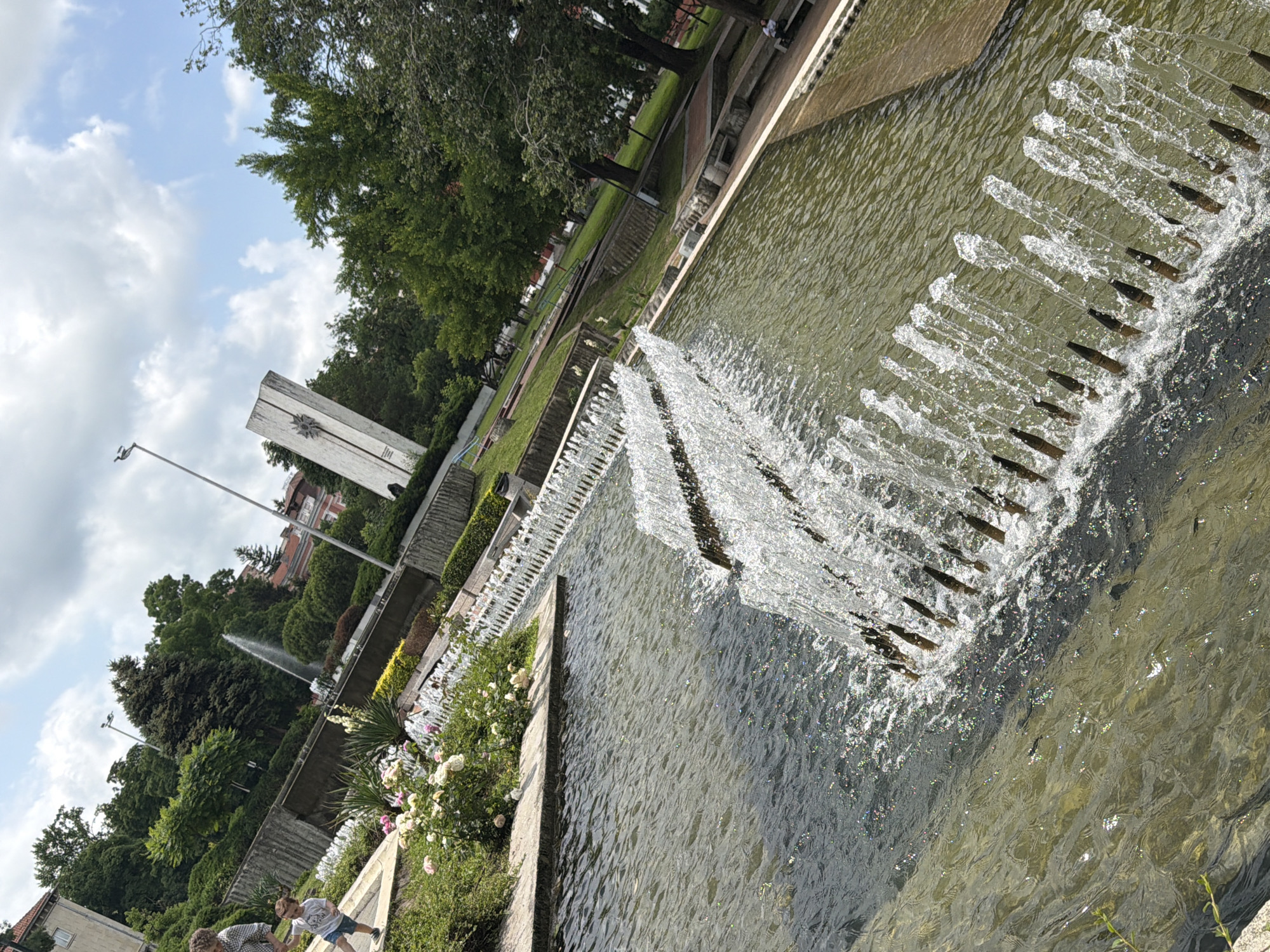 This image captures an outdoor park or public garden scene, likely under a partly cloudy sky during daylight hours. The central feature is a multi-tiered water fountain. The lower tier of the fountain displays numerous individual jets spraying water upwards, creating a lively display, with sunlight causing bright glints and sparkles on the moving water. Water also cascades down from an upper level into the main pool.
The surroundings are lush with vegetation, including abundant green trees, bushes, and flowering plants, notably white and pink roses, lining the fountain and pathways. In the background, a tall, white concrete monument or structure with a distinct star-like emblem or carving is prominent. Further back, buildings with reddish roofs are visible. The sky is partly cloudy with patches of blue.
Two individuals, appearing to be an adult and a child, are partially visible in the lower left foreground, near the flowering plants, suggesting a recreational visit to the park. The scene conveys a peaceful and pleasant atmosphere. Faint, illegible text or markings can be observed on the side of the white monument below the star emblem. The setting aligns with public spaces in Pleven, Bulgaria.