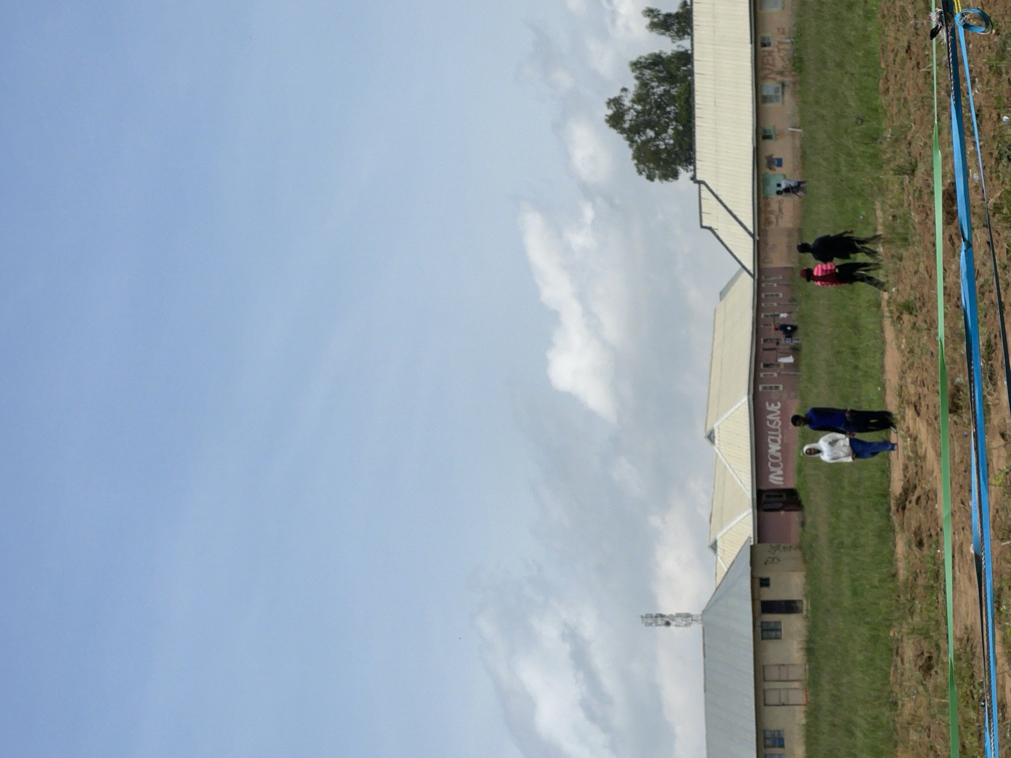 The image depicts an outdoor scene on what appears to be a grassy field adjacent to a building with a white, sloped roof and beige walls. The building has windows and doors visible. In the foreground, there are several people walking or standing. Two individuals are walking together in the middle ground, one wearing a black shirt and the other a red and black patterned jacket. Further down the field, another pair of people are standing, one in a blue shirt and the other in a white hooded sweatshirt. A cell tower is visible in the distance. There are also several colored ropes or ribbons stretched vertically along the right side of the frame, appearing to be part of some sort of marking or boundary. The sky is light blue with some scattered white clouds, suggesting daytime. The grass is green and appears well-maintained. There is some text visible on the side of the building: "INCONCLUSIVE" is written in large, red, graffiti-style letters. The overall impression is of a casual outdoor gathering or event in a rural or semi-rural setting in Durumi, Nigeria.