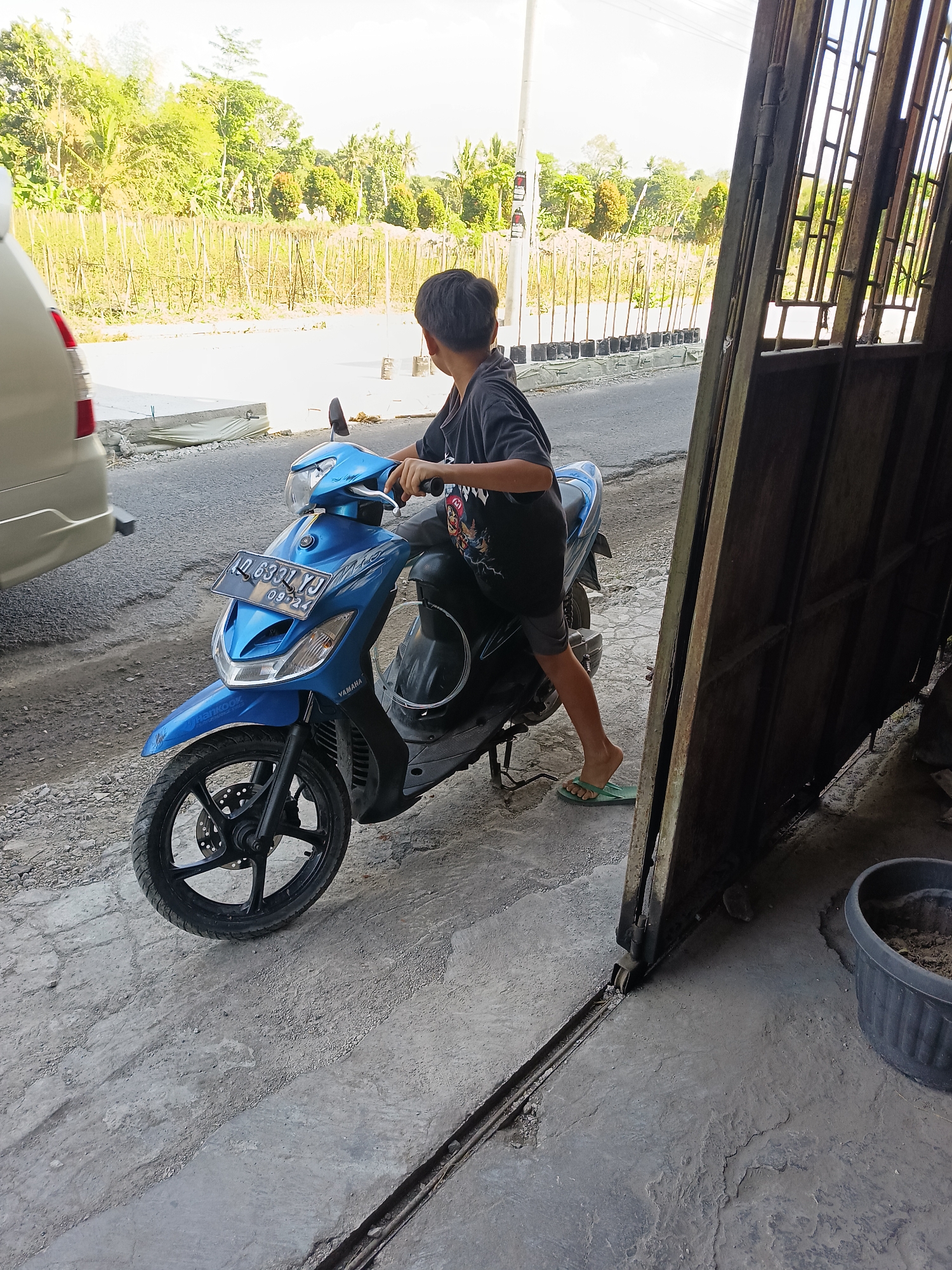 A young boy is sitting on a blue Yamaha scooter parked on a concrete surface. He is wearing a dark t-shirt and shorts. The scooter has a license plate that reads "AD 6331 YJ 09-24". The scene appears to be in Sukorini, Indonesia, as indicated by the Indonesian license plate and the surrounding tropical vegetation. In the background, there is a fence made of bamboo poles and some buildings. To the left of the scooter, part of a beige car is visible. To the right, a metal gate and a large black plastic pot are present. The lighting suggests it is daytime.