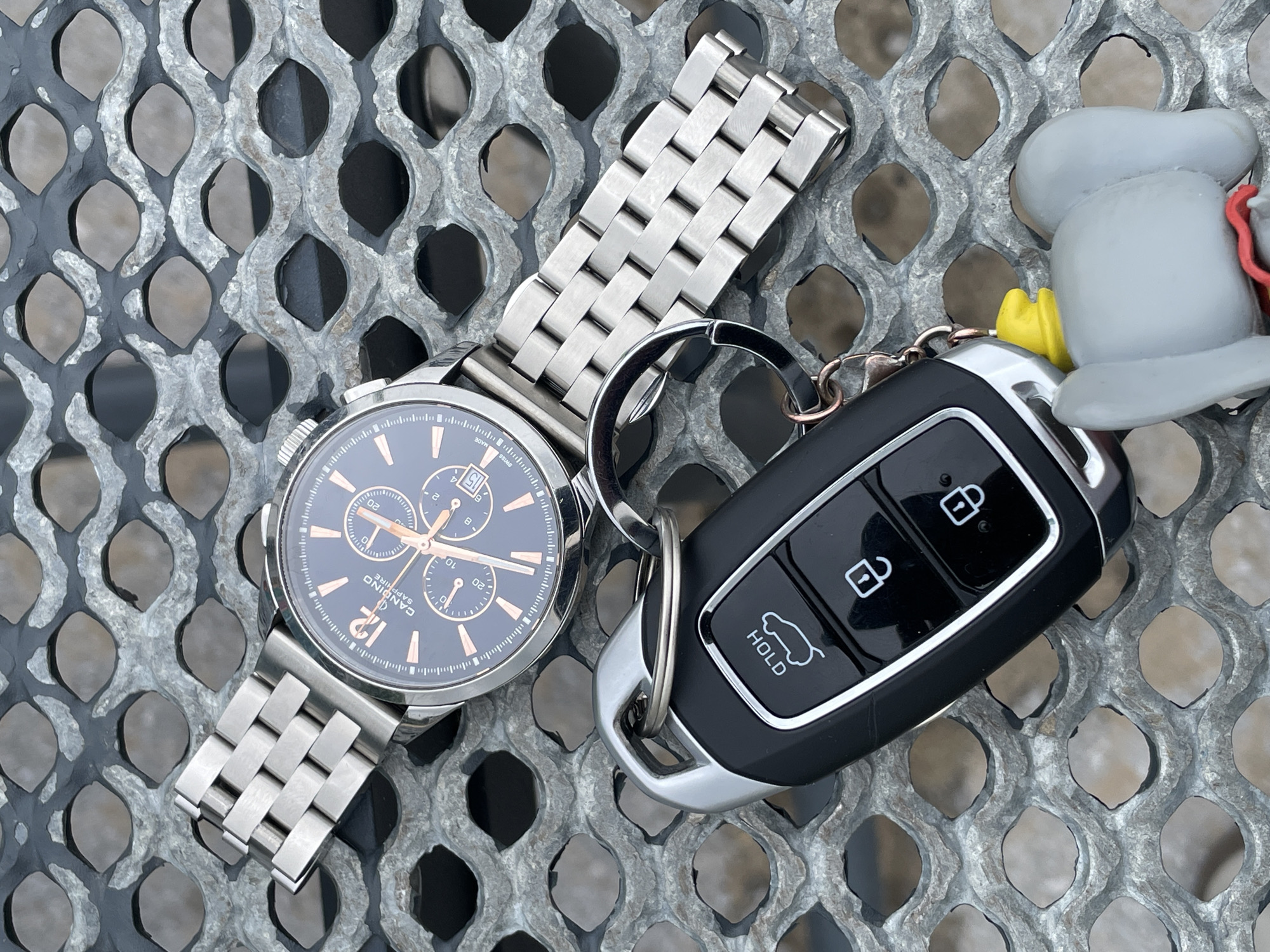 On a gray, metal, lattice-patterned surface, a wristwatch and car keys are arranged. The wristwatch has a silver metal band and a dark face with light-colored hands and numerical markings. The car keys are attached to a keyring and include a black fob with silver accents and labeled buttons. A small, gray, cartoon-like figurine is attached to the keyring. The location is unspecified.