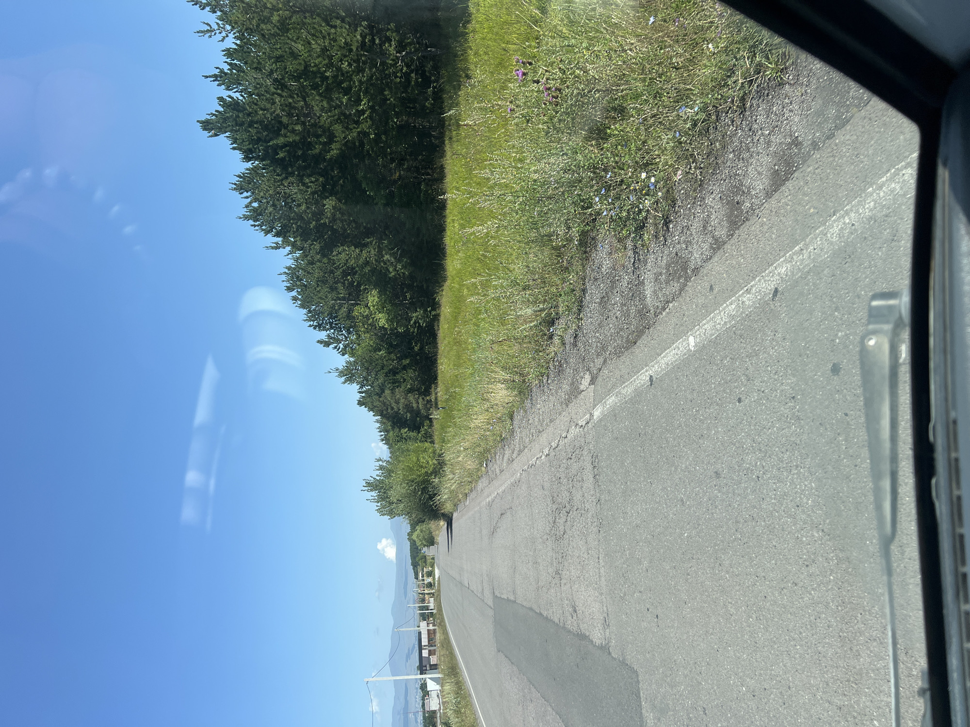 The image depicts a rural road leading into the city of Aragva, Georgia. The road is asphalt, with visible patches and cracks, suggesting it is well-traveled but not immaculately maintained. Power lines and poles are visible along the left side of the road, indicating the presence of infrastructure. In the distance, a few low-rise buildings and unfinished construction projects are discernible. The right side of the road is bordered by a grassy field with scattered wildflowers, beyond which lies a dense forest of pine trees. The sky is a clear blue with a few wispy clouds. The scene appears to be captured from inside a vehicle, as evidenced by the windshield wipers and the corner of the vehicle's interior visible at the bottom of the frame. The overall impression is one of a peaceful, somewhat undeveloped, semi-urban landscape on a sunny day. There are no people or animals clearly visible in the image.