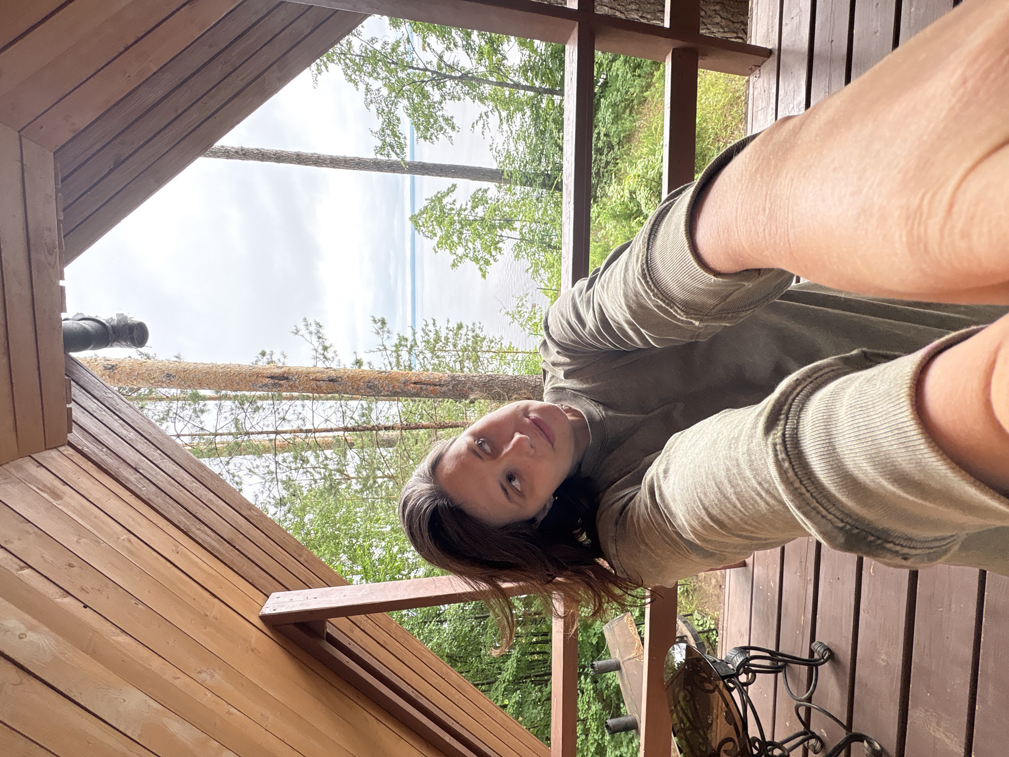 The image captures a woman taking a selfie on a wooden porch overlooking a body of water and lush green trees. The woman, with dark hair, is wearing a long-sleeved, olive-green shirt. Her left arm is extended towards the camera, and she is looking to her right. The porch is constructed of wooden planks and has a wooden railing. Behind the railing, a wide expanse of water is visible, with trees lining the shore and extending into the distance. The sky is overcast. In the foreground, to the left, a portion of a dark, ornate table with a glass top is visible. The overall setting suggests a tranquil natural environment. The location is identified as Kostovaty, Russia.