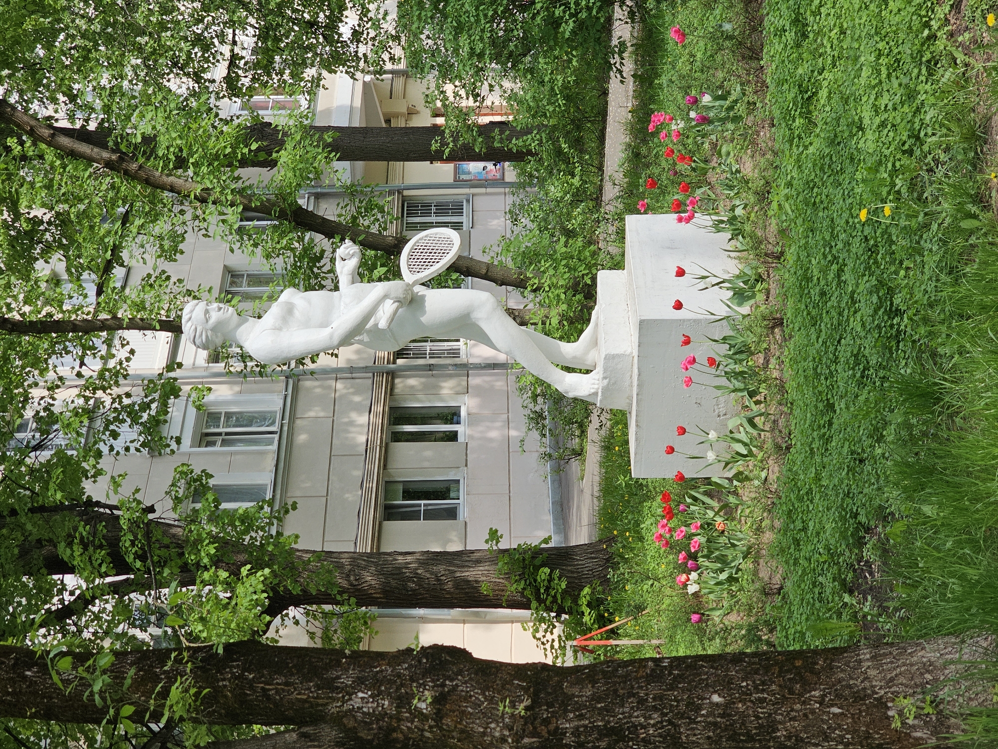 The image shows a white statue of a tennis player in a park setting in Moscow, Russia. The statue is reclining on a white pedestal, holding a tennis racket. Red and pink tulips grow around the base of the pedestal. Behind the statue is a residential building with white windows. Trees are visible in the foreground, partially obscuring the view. The setting is green and lush with grass and foliage.
