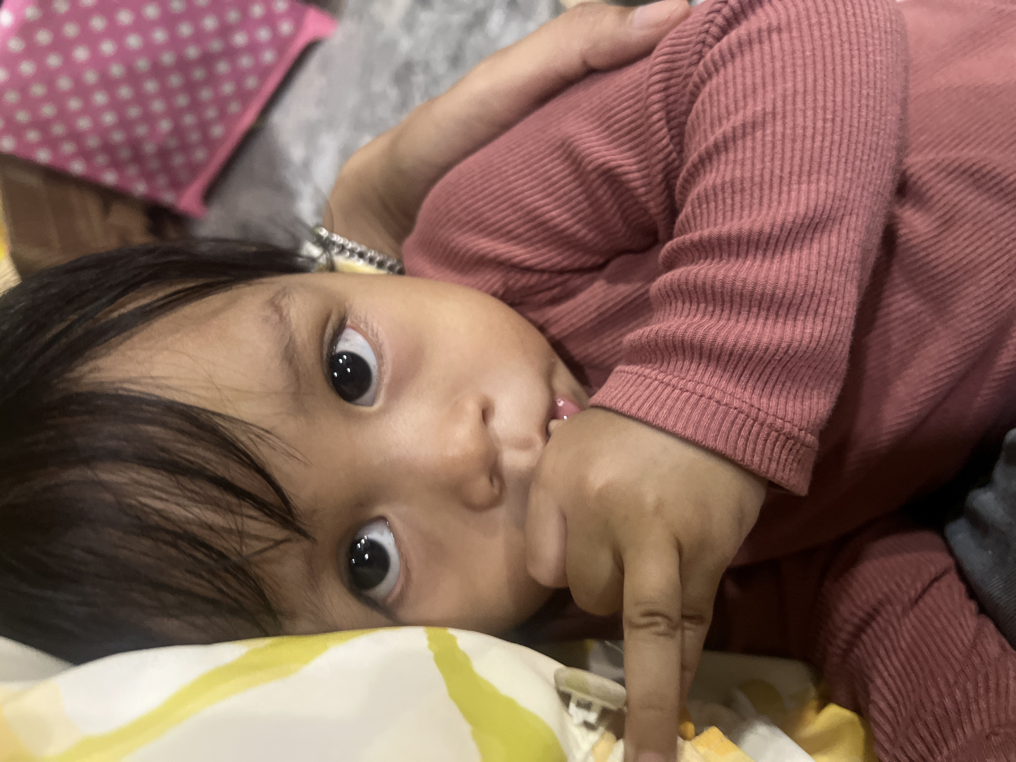 A close-up shot focuses on the face of a young child, who appears to be an infant or toddler. The child has dark hair, dark eyes, and is looking directly at the camera with a slightly curious expression. Their right hand is raised to their mouth, with fingers placed inside. The child is wearing a dark pink, ribbed long-sleeved shirt. To the left of the child's face, a portion of a yellow and white patterned fabric is visible, possibly a blanket or pillow. In the background, a blurred pink object with white polka dots is discernible, and to the right, an adult's arm is visible, lightly embracing the child. The overall setting appears to be indoors, with a neutral-colored, textured surface in the background, suggestive of a carpet or rug. The image was taken in Wedarijaksa, Indonesia.