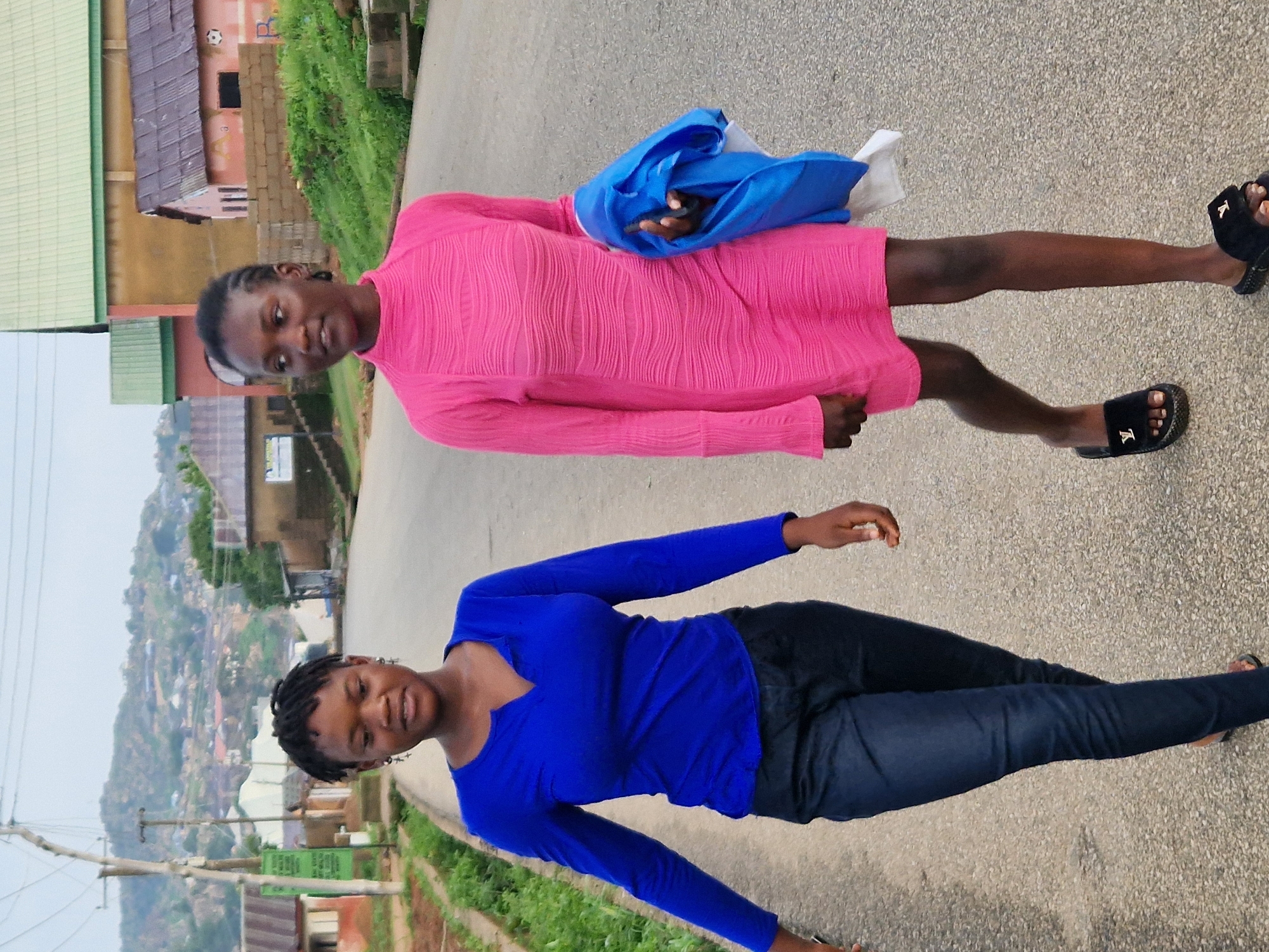 Two young Black females are walking on an asphalt road in Kabong, Nigeria. The female on the left wears a royal blue long-sleeved shirt and dark wash jeans. She has braided hair and is wearing cross-shaped earrings. The female on the right wears a pink, textured, long-sleeved dress and black slides. She is carrying a blue cloth in her right hand. The road is lined with green vegetation and a few buildings. In the background, a hilly landscape dotted with houses is visible under a hazy sky. The scene appears to be a residential area.