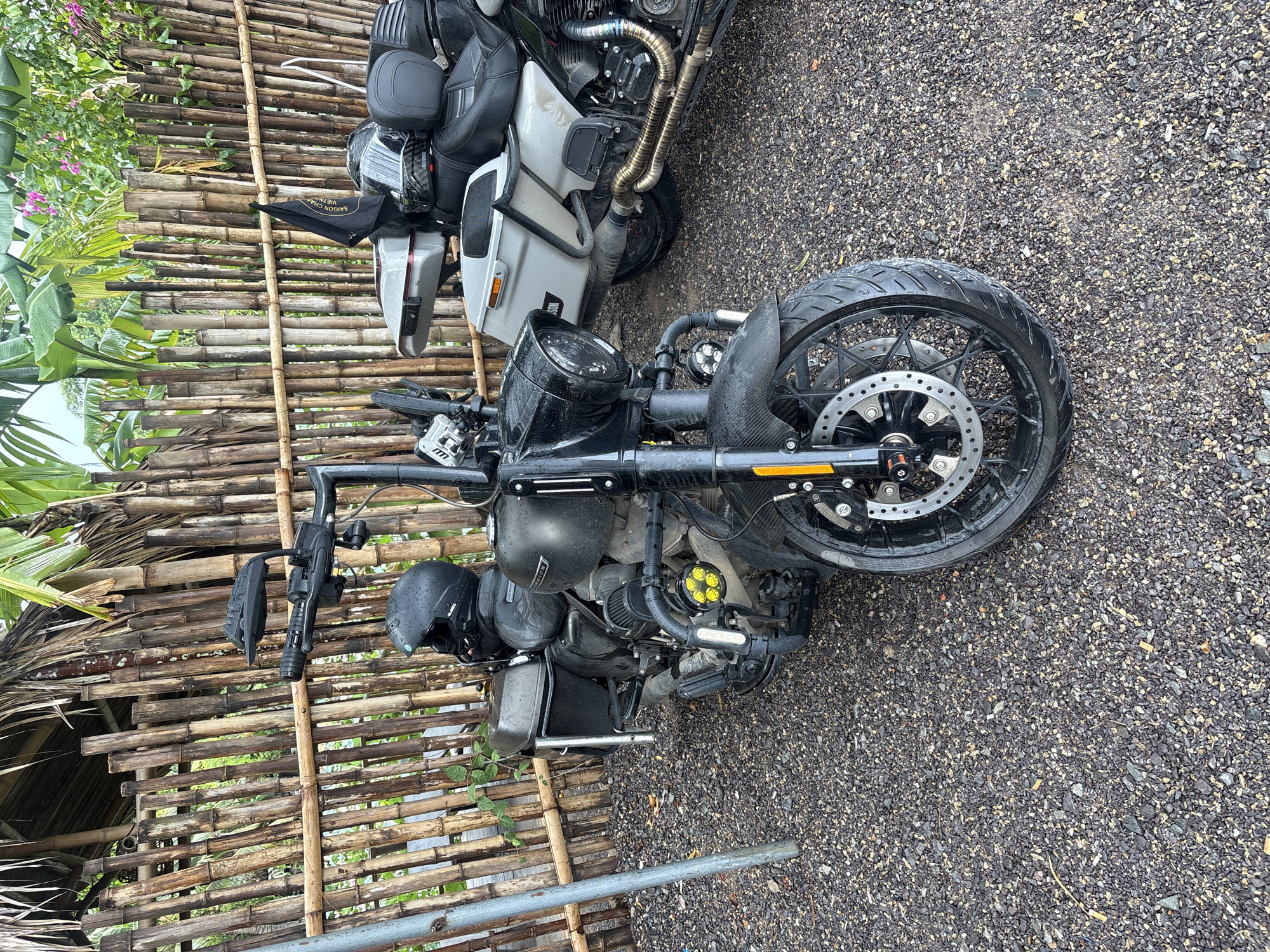 The image shows two motorcycles parked on a gravel surface. One motorcycle is black, with a front wheel prominently displayed. A helmet and other accessories are attached to the motorcycle. A bamboo fence runs along the side of the motorcycles, with vegetation visible beyond the fence. The second motorcycle is grey, positioned behind the black one, and appears to be partially loaded with luggage. Based on the metadata, the image was taken in Nam Nha Trang Ward, Vietnam.