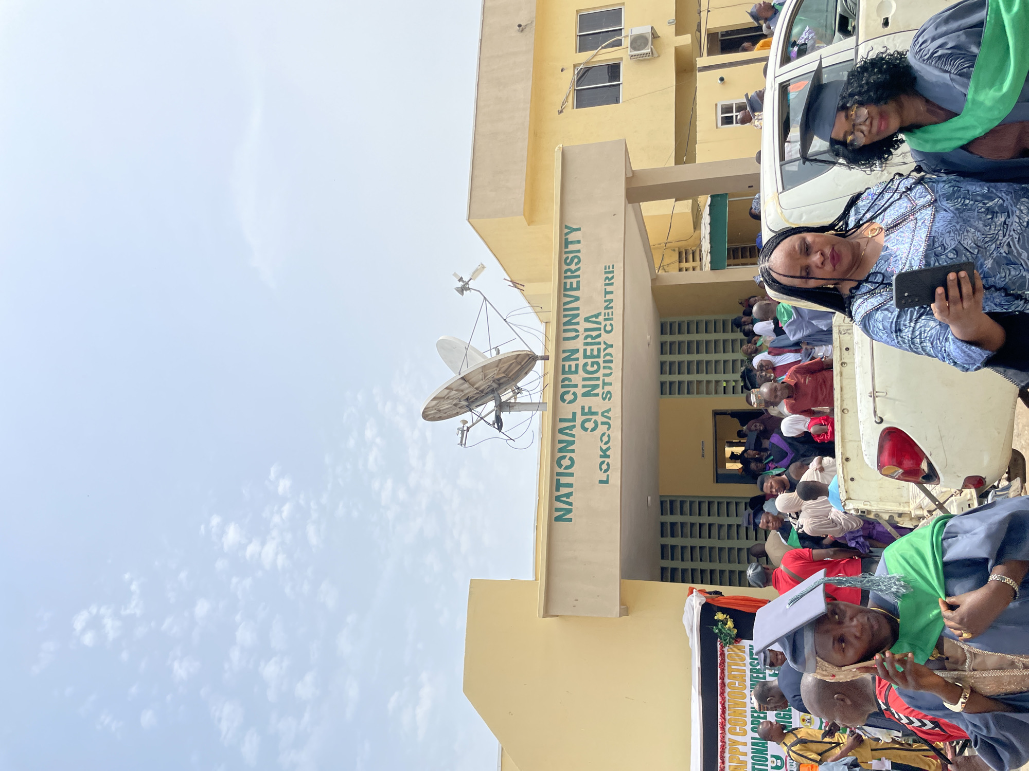 The image shows the National Open University of Nigeria, Lokoja Study Centre. The building is a light yellow color with a beige-colored entrance. A satellite dish is mounted on the building. Several people are visible, many of whom are wearing graduation gowns and caps, suggesting a graduation ceremony or event. Some individuals are holding phones. The scene is crowded with attendees. The image orientation is not horizontal. The sky is pale blue with some clouds. A vehicle is near the entrance of the building. There is a banner in front, though the text is partially obscured.