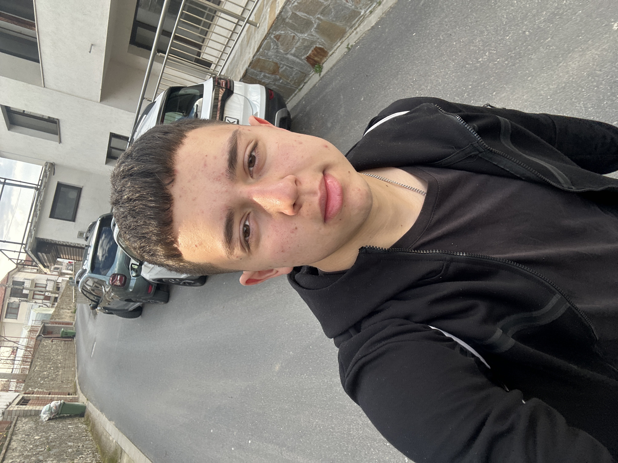 A selfie of a young man with short, dark hair wearing a black T-shirt and a black zip-up hoodie with white stripes. He is wearing a silver chain. The photograph is taken outdoors on a paved street in what appears to be a residential area in Zlatograd, Bulgaria. Several cars are parked along the street, and buildings are visible in the background. There are a couple of trash cans in the background as well. The buildings are a mix of older, stone-walled structures and more modern, multi-story buildings.
