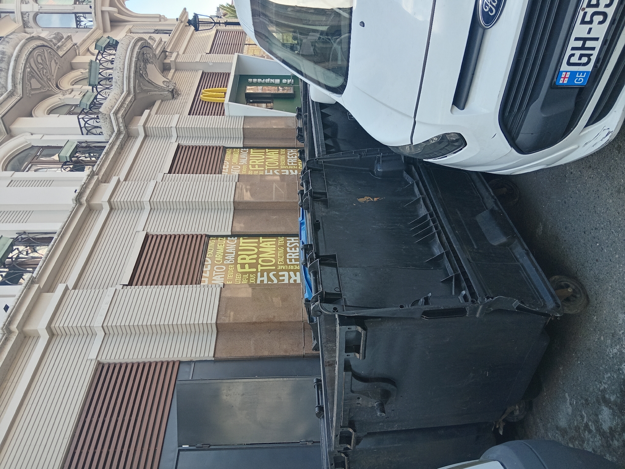 A daytime shot shows a white Ford van parked next to black trash bins in the city of Batumi, Georgia. The van's front end, including a license plate marked "GH-55" with the Georgian flag, is visible. The building behind the bins features a McDonald's Express sign in green and white. A sign on the building facade displays words such as "EXCITING," "CARAMEUZ," "TENDER," "BALANCE," "FRUIT," "TOMATO," and "FRESH." The building itself features an elaborate facade with architectural details, including balconies with iron railings.