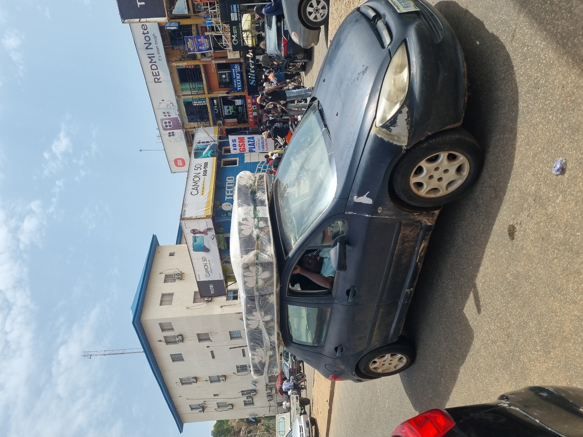 The image captures a street scene in Jos, Nigeria, characterized by commercial activity and a mix of buildings and infrastructure. In the foreground, a dark blue car is noticeably overloaded with a stack of mattresses secured to its roof. The driver can be seen inside the car. The car is on a paved street surrounded by other vehicles, including cars and motorcycles. In the background, a multi-story building houses various businesses, as evidenced by the numerous signs and advertisements. Prominent advertisements feature brands like TECNO and REDMI Note. The building appears to be a commercial plaza or shopping complex. Numerous people are visible on the street, suggesting a bustling urban environment.