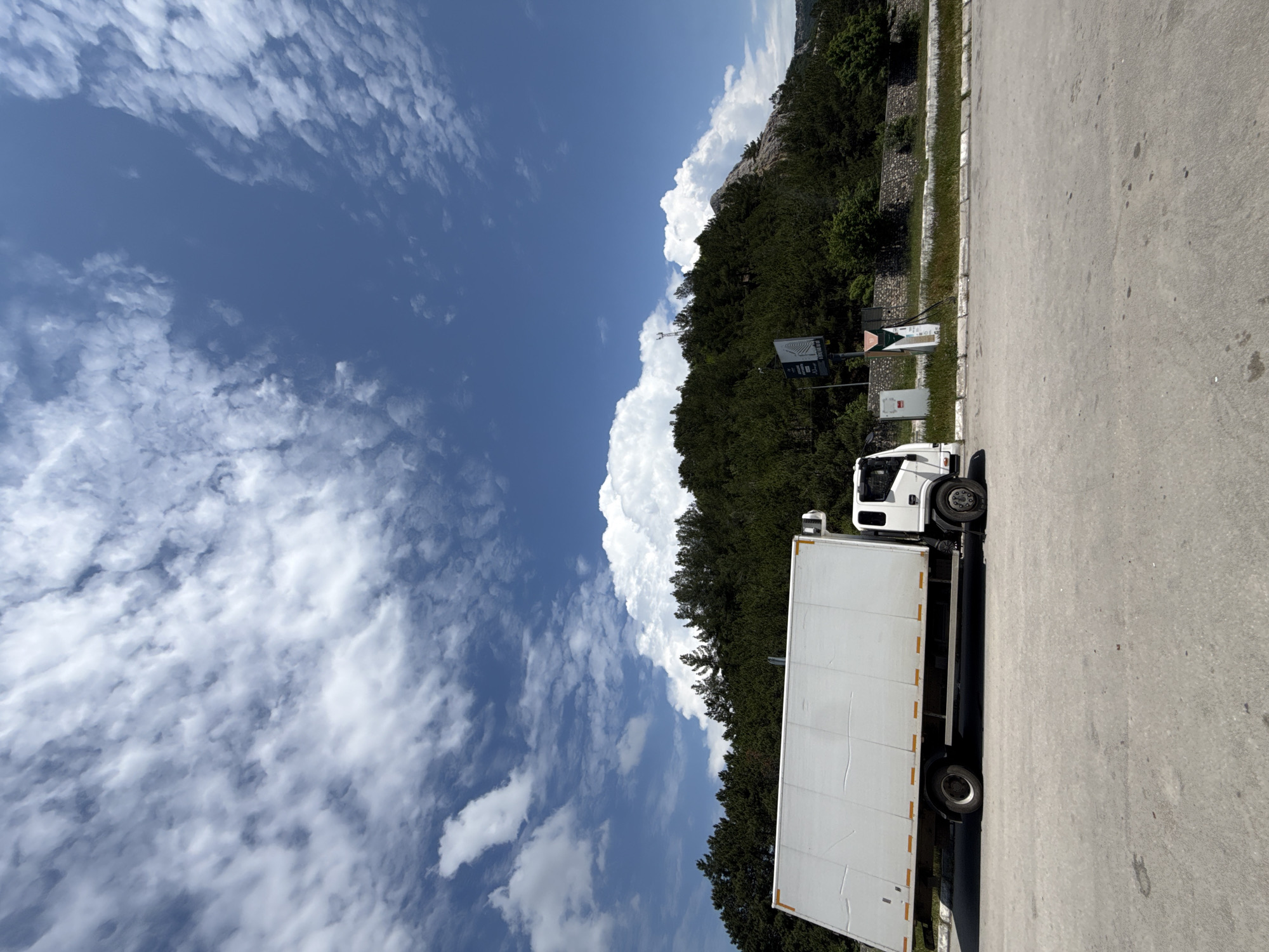 A white refrigerated truck is parked on a paved surface, possibly a roadside. The truck is oriented horizontally in the frame, with its front facing right. The background features a densely forested hillside under a bright blue sky with scattered white clouds. A rocky outcrop is visible on the right side of the hillside. In the middle ground, a stone wall borders the forested area, and several utility boxes and poles are present. The scene is static and captures a daytime view.