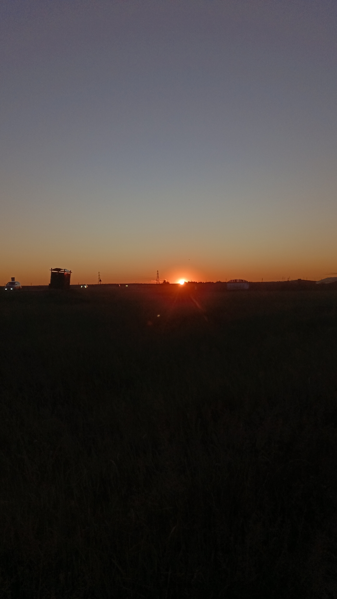 The image depicts a sunset over a rural landscape, likely on the outskirts of Angarsk, Russia. The sun is low on the horizon, casting a warm, orange glow across the sky and creating a lens flare effect. In the foreground, a dark, grassy field stretches across the frame. In the mid-ground, several structures are visible: a large, dark, box-like structure with a roof, resembling a hunting blind or observation post, is on the left. To the right of this, several vehicles with illuminated headlights are driving on a road. Further to the right, there are power transmission towers and some distant buildings. The sky transitions from a deep blue at the top to a vibrant orange near the horizon.