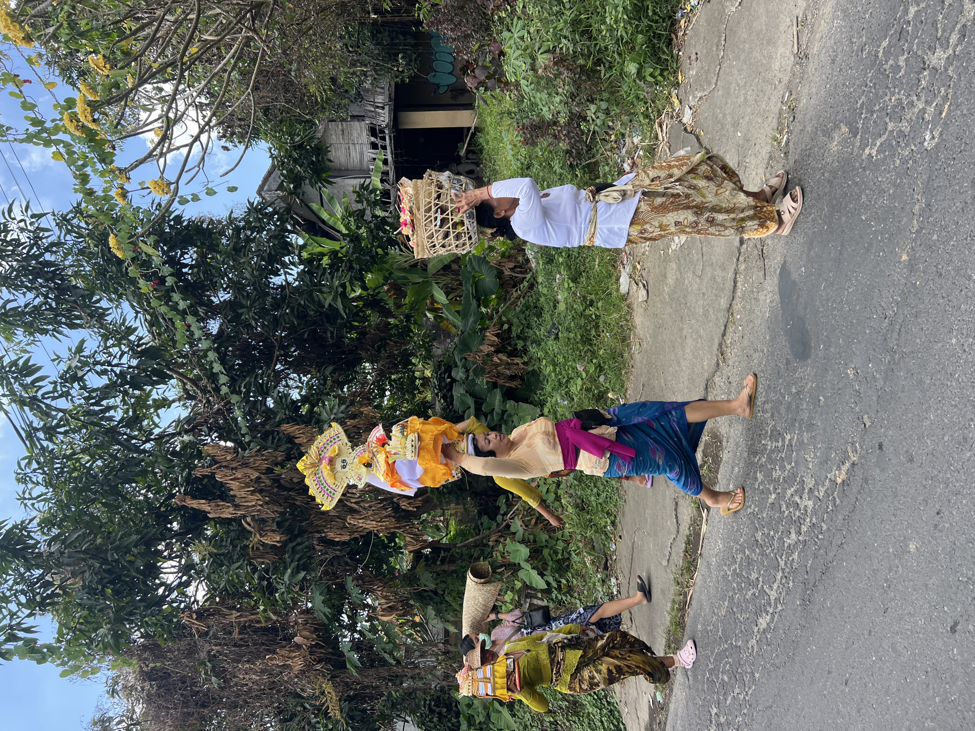 Four individuals are walking on a paved road in what appears to be a rural area near a building, with lush green foliage and trees surrounding the scene. The individuals are carrying items on their heads. The first person on the left, dressed in a yellow top and patterned skirt, carries a yellow tray with a decorative offering. Behind her, a person in a pink top and a face mask carries a woven mat. The third person, in a gold-colored top and a blue sarong, carries a tall, ornate offering on their head. The fourth person on the right, wearing a white top and a patterned skirt, carries a basket filled with flowers and fruit on their head. The environment suggests a tropical or sub-tropical climate, with the building in the background appearing somewhat dilapidated. The road surface is asphalt with visible cracks and patches. The sky is blue with scattered clouds. The overall impression is of a procession or a ritualistic activity taking place outdoors.