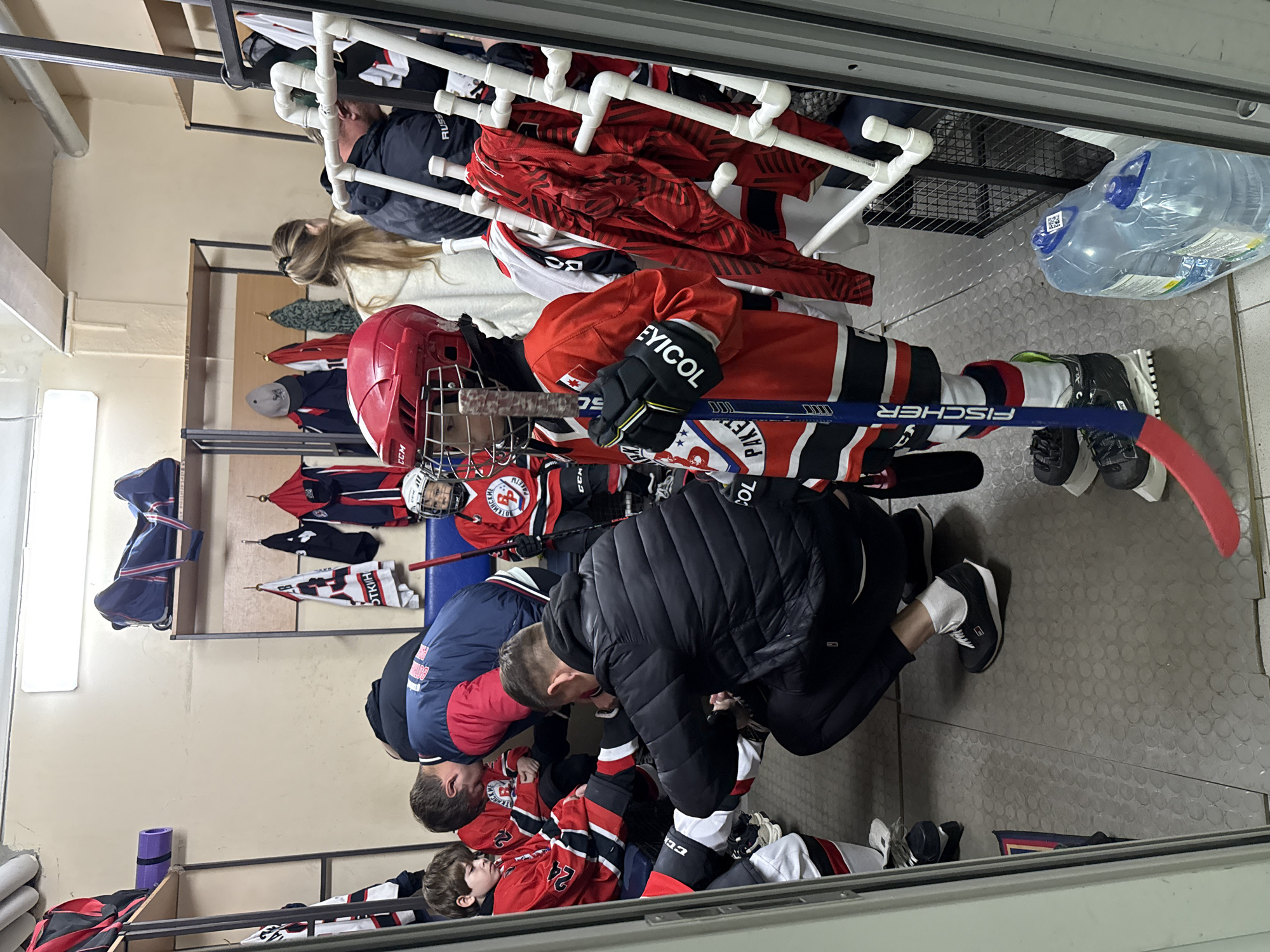 **Location:** Izhevsk, Russia, likely a locker room or dressing room.
**People:** Multiple hockey players (children) are present, wearing red and black hockey jerseys, helmets, and equipment. Adults (likely coaches or parents) are helping the children with their equipment.
**Subjects:** Hockey players are the primary subjects.
**Objects:** Hockey sticks, hockey skates, red and black jerseys, helmets, gloves, water bottle.
**Actions:** Adults are helping children with their hockey equipment, such as tying skates. Players are getting ready for a game.
**Interactions:** Adults are interacting with children, providing assistance. Players are interacting with each other.
**Environment:** The environment is a locker room with benches, shelves, and hanging racks for equipment. The room is brightly lit.