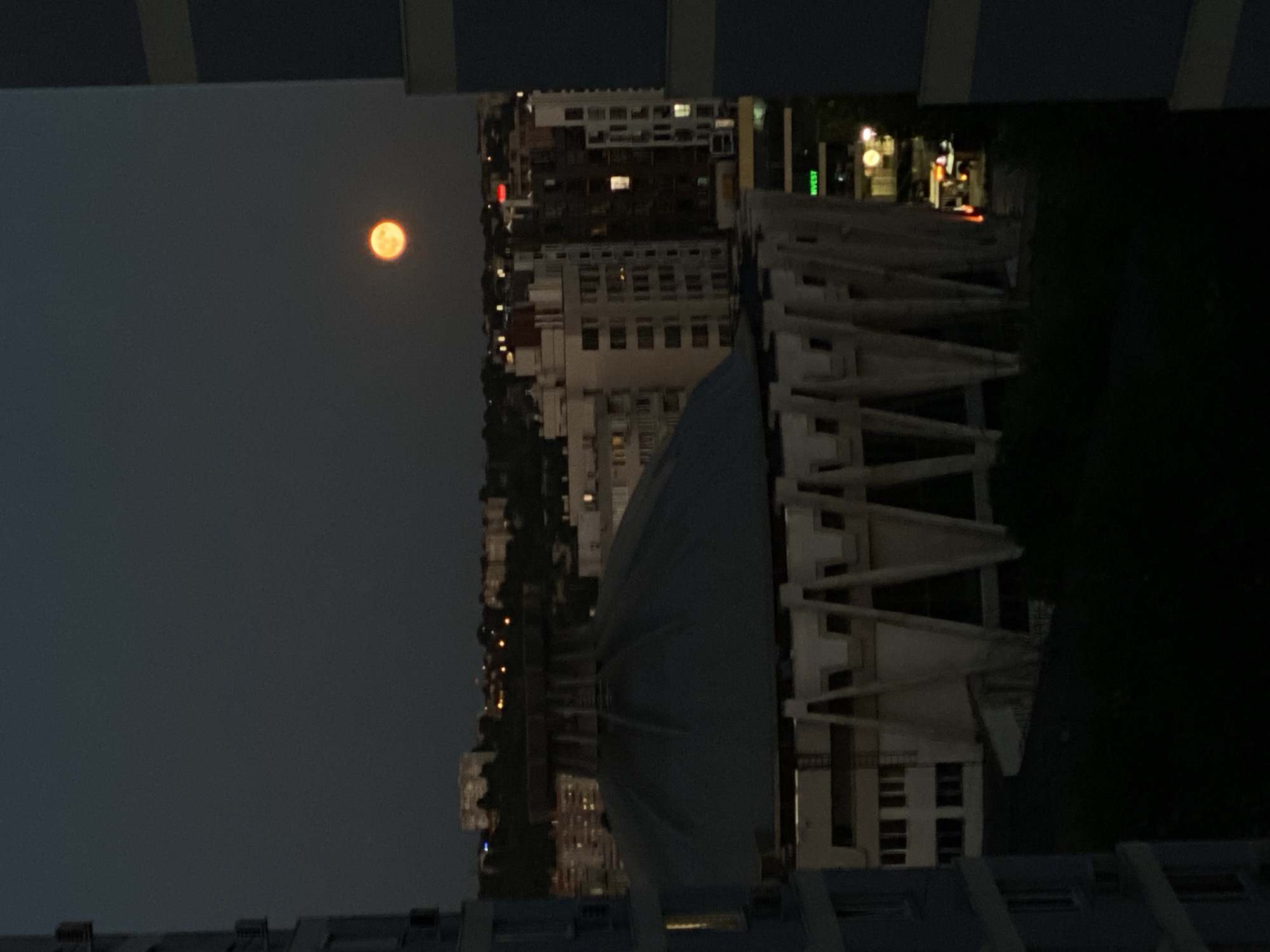 The image, taken from a high vantage point within the city of Chișinău, Moldova, captures a twilight cityscape dominated by a large, domed structure in the foreground. The moon, a vibrant orange orb, hangs high in the dark sky. In the middle ground, a series of multi-story buildings with illuminated windows form the urban fabric, interspersed with trees. The foreground is partially obscured by a dark, unlit area, suggesting the presence of a lower building or ground level. The domed building, with its distinctive angular supports, appears to be a public building, possibly a sports arena or cultural center, and is characteristic of Soviet-era architecture found in Chișinău. The overall impression is one of urban stillness as night begins to fall, punctuated by the natural spectacle of the moon.