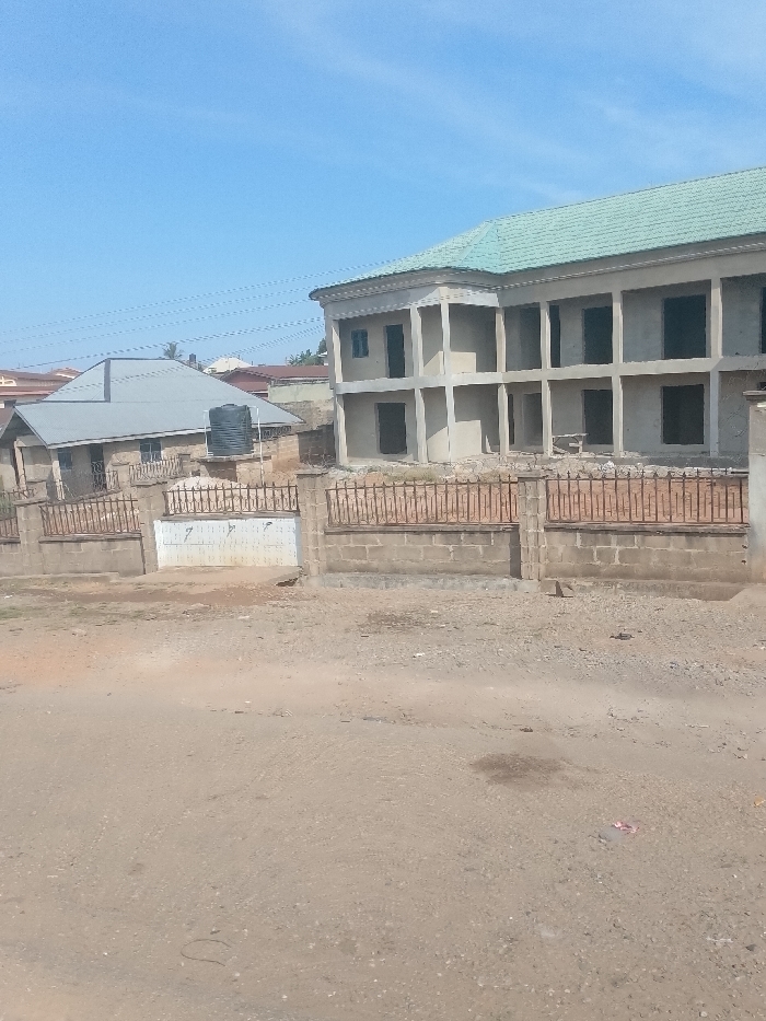 The image displays an urban scene in Ayegbaju, Nigeria, under a clear blue sky. In the foreground, an unpaved dirt and gravel road extends across the frame. A low boundary wall, constructed from concrete blocks and topped with dark metal wrought iron fencing, runs through the midground. Integrated into this wall is a light-colored, double-leaf metal gate. Behind the fence, to the right, stands a multi-story building currently under construction, identified by its exposed concrete structure, numerous unfinished window and door openings, and a light green corrugated metal roof. To the left of this, a smaller, completed single-story building with a gray corrugated metal roof is visible, featuring a black water storage tank elevated on a stand beside it. No individuals are present in the scene.