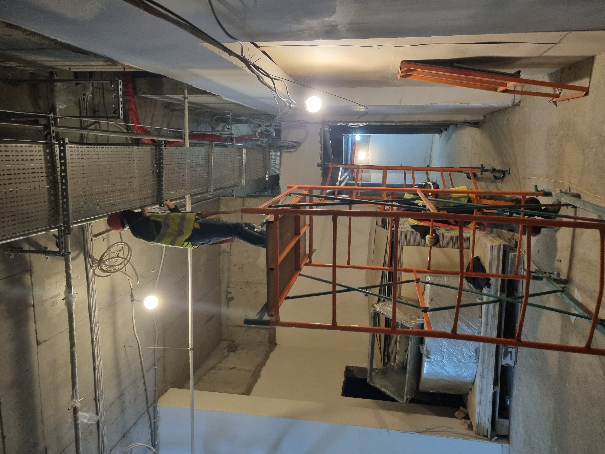 A construction worker wearing a hard hat and safety vest is working on scaffolding inside a building under construction. The location is likely Sochi, Russia. The scaffolding is orange, and the worker is standing on the upper level. The building appears to be a concrete structure with electrical wiring and conduits visible on the ceiling and walls. There are metal cable trays mounted on the ceiling. There are also building materials stacked and wrapped in plastic near the scaffolding. A light fixture is suspended from the ceiling.