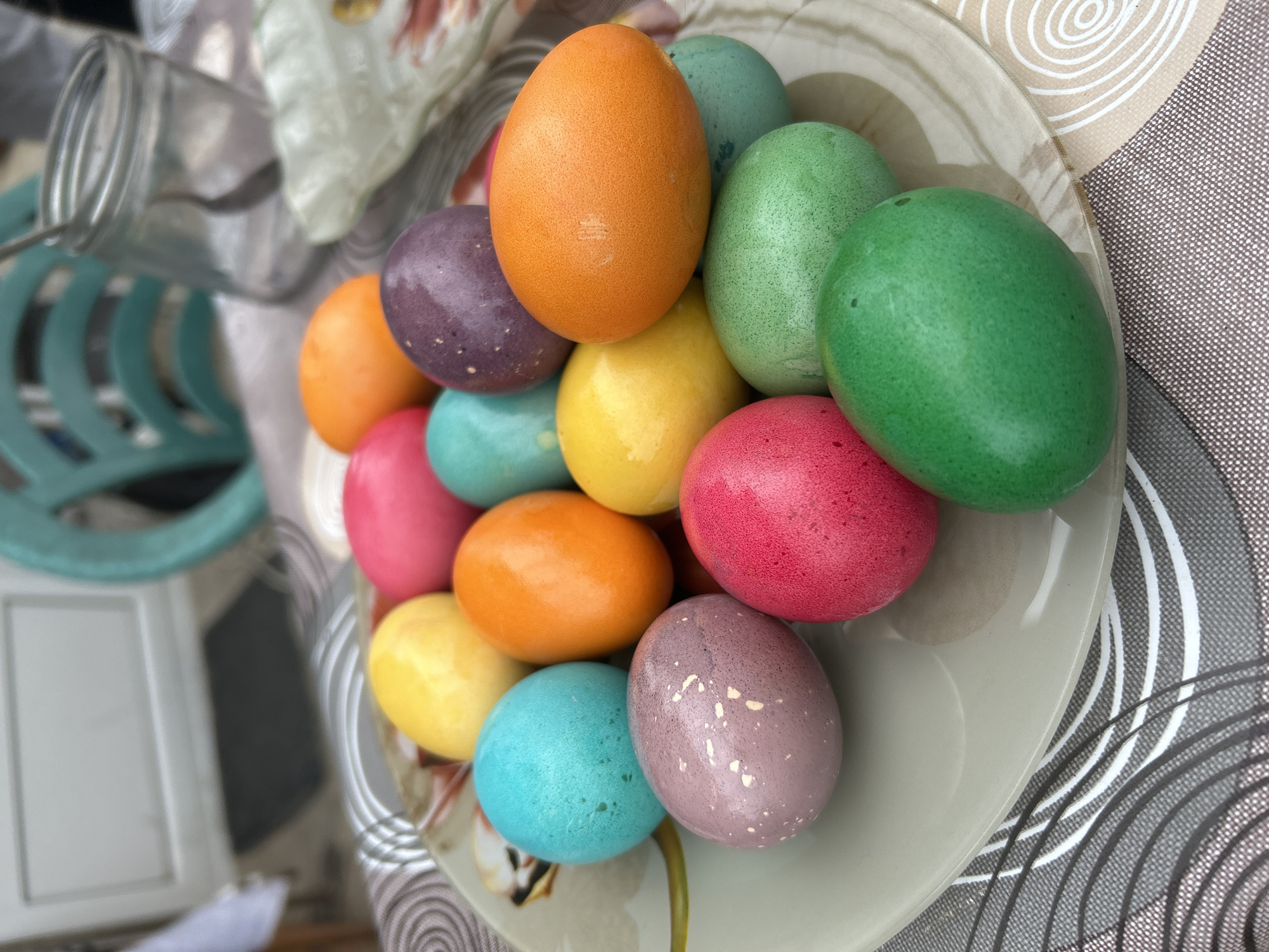 A top-down shot shows a plate piled high with brightly colored Easter eggs, mostly in shades of orange, purple, yellow, blue, red, and green. The plate sits on a tablecloth with a circular pattern in shades of gray, black, and white. A glass jar is slightly out of focus in the left of the image. The image does not provide enough information to ascertain if it was taken in Zlati Voyvoda, Bulgaria. There is no information provided in this image that can determine if people are in this scene or interacting.