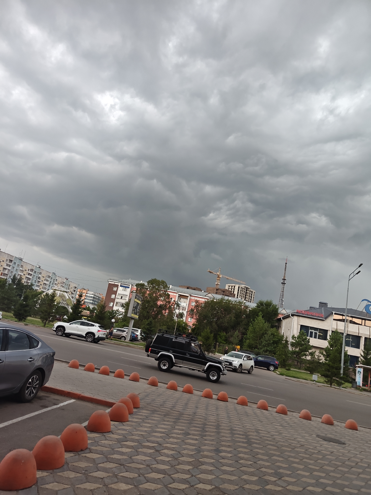 The image depicts an overcast sky with heavy, dark clouds over a city street scene in Petropavl, Kazakhstan. In the foreground, a grey car is parked on the left, with a row of orange, dome-shaped bollards separating the sidewalk from the street. The street has multiple lanes of traffic. Several cars are visible on the road, including a white SUV, a black SUV, and other vehicles. In the middle ground, there are trees and several multi-story buildings, some of which appear to be residential apartments and commercial establishments. Construction cranes are visible in the distance, indicating ongoing development. The overall atmosphere is somber due to the dark clouds.