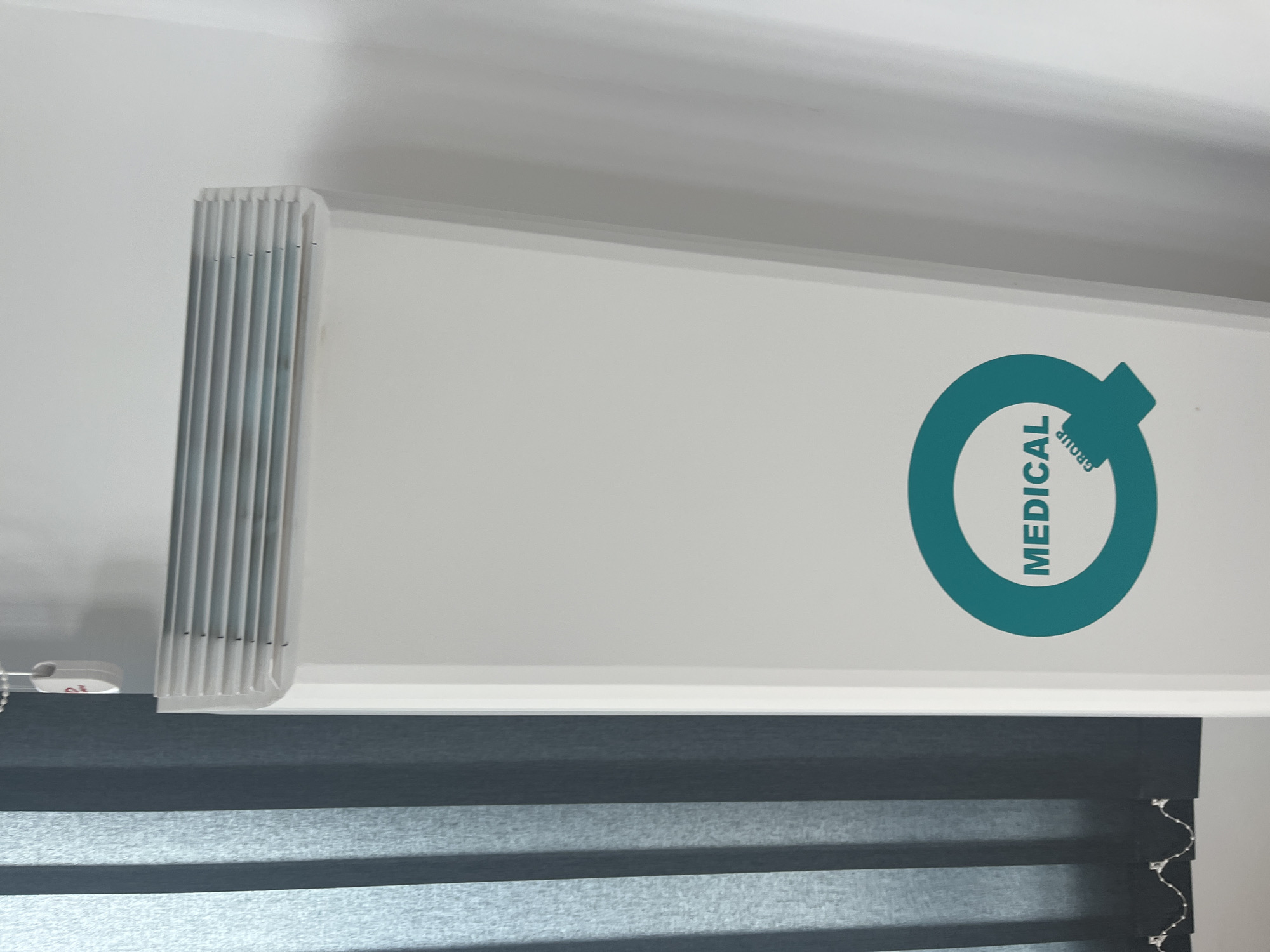 The image shows the top portion of a window with a partially drawn blind, set against a plain white wall. The blind is a dark gray and white striped pattern. Above the blind is a white valance or headrail bearing a teal-colored logo that includes the word "MEDICAL" oriented vertically. To the left of the valance is a series of vertical slats, possibly part of the blind mechanism. The image was taken indoors. Without additional context, the location in Plovdiv, Bulgaria is not determinable.