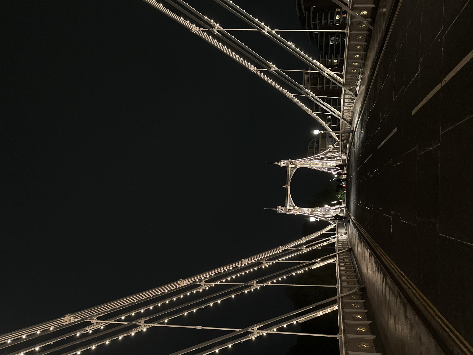 The image is taken from a low perspective, looking up and down the illuminated Albert Bridge in London, United Kingdom at night. The bridge's suspension structure is adorned with strings of lights, emphasizing its architecture. A dark asphalt road runs alongside, marked with faint yellow lines. In the distance, a section of the bridge's towers is visible, also lit up. The sky is a deep, almost black, night sky.