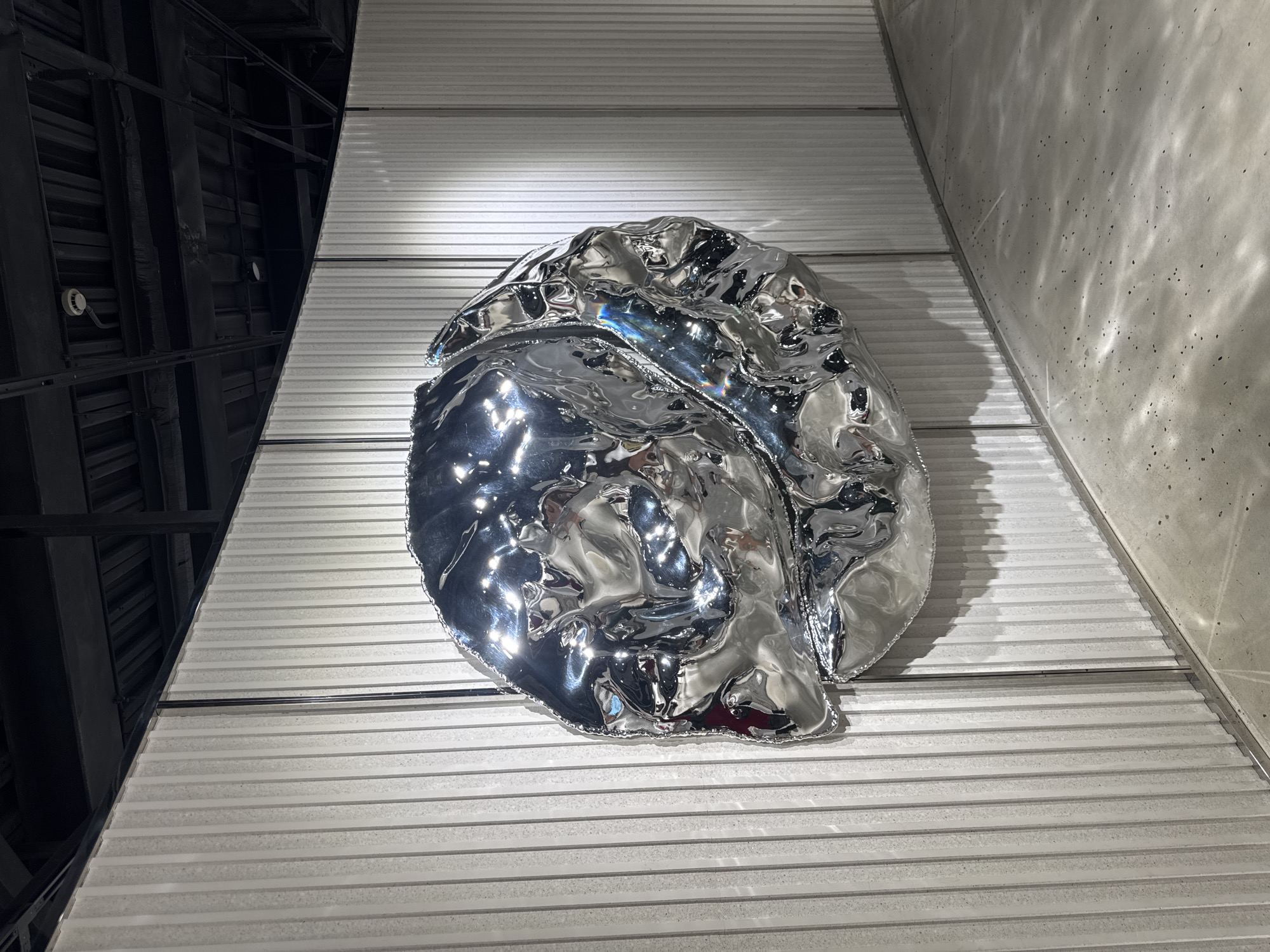 A large, crumpled, metallic sculpture is mounted on a curved, ribbed white wall in an indoor exhibition space. The sculpture is highly reflective, showing distorted images of its surroundings. The wall appears to be made of textured panels. The floor is a polished concrete surface with scattered dark aggregate. The ceiling above is dark with visible industrial elements like ducts and lighting fixtures. The overall impression is of a modern art installation. The location is confirmed to be the X Museum in Shanghai, China. There are no people visible in the image.
