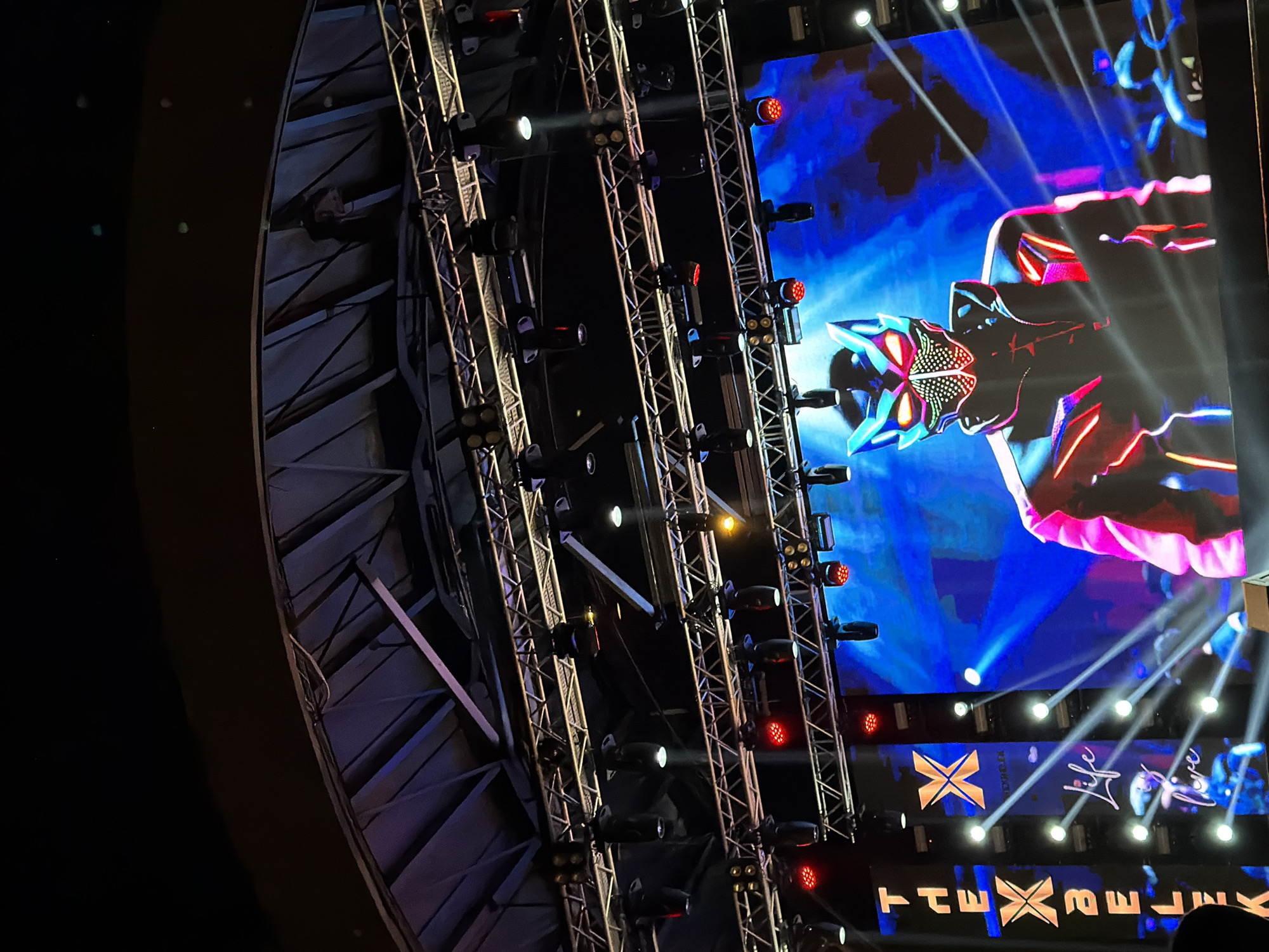 The image captures a vibrant stage setup at what appears to be a concert or large entertainment event, taking place at night or indoors. The scene is dominated by multiple metallic truss structures, densely packed with numerous stage lights. Many lights emit powerful white beams cutting through the dark atmosphere, while others glow with intense red light.
To the right, a large LED screen displays dynamic, colorful graphics. A prominent feature on the screen is a stylized, possibly robotic or anime-inspired character's head and upper body, rendered in striking blues, pinks, and reds with sharp lines and glowing elements. Below this character, visible text on the screen spells out "THE XPERIENCE" (or "THE XPRNC" with a stylized 'E'), alongside a distinctive "X" logo. Further to the right, the words "Life" and "Live" are also displayed in an elegant, script-like font.
On the far left, a segment of a large, dark, and curved architectural structure is partially visible, suggesting the interior of a substantial venue or an elaborate stage design. The perspective is from below, looking up towards the stage and lighting rig, conveying the impressive scale and high-tech nature of the production. The overall impression is one of energetic spectacle, typical of a major visual and audio performance.