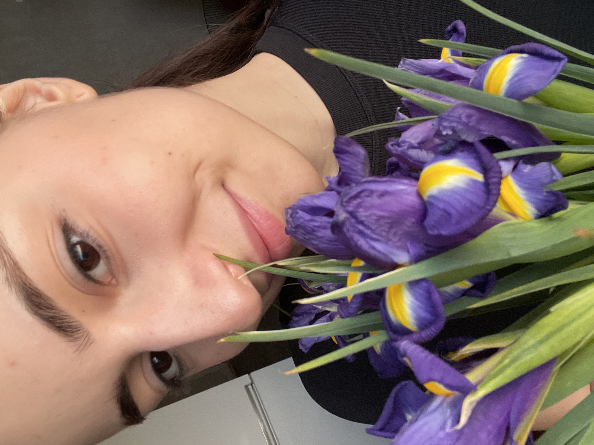 The image portrays an adult female subject in a close-up, head-and-shoulders view, situated indoors in the city of Moscow, Russia. She has a light complexion, dark hair pulled back, and dark eyes, looking forward with a subtle smile. She is wearing a black, ribbed, long-sleeved crew-neck shirt. A bouquet of purple irises, accented with distinct yellow and white markings on their petals and interspersed with green foliage, is held close to her face, partially obscuring her nose and mouth. The background is out of focus, showing a light-colored vertical surface on the left and a darker, indistinct area on the right.