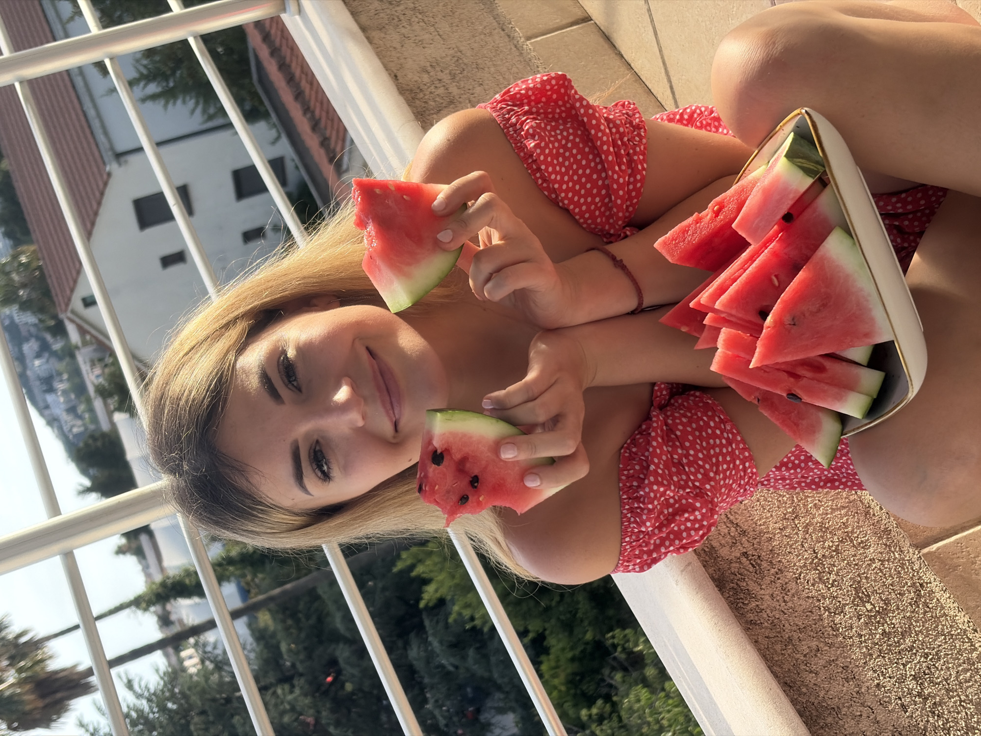 A young woman with long, blonde hair and a radiant smile is captured in a high-angle shot on a sunny day. She is wearing a red and white polka-dotted top. The woman is holding two slices of watermelon, one in each hand, and appears to be enjoying them. To her right, a white bowl filled with several triangular slices of watermelon rests on her leg. Her eyes are open, and she is looking directly at the camera with a pleasant expression.
The setting appears to be a balcony or outdoor patio, indicated by the white railing in the foreground and the textured, light-brown ground beneath her. In the background, beyond the railing, residential buildings with tiled roofs and lush green trees are visible, suggesting a scenic, possibly coastal or hilly, location like Budva, Montenegro. The warm sunlight hitting the scene suggests it is either morning or late afternoon. No visible text is present in the image. The overall mood is relaxed and joyful, capturing a moment of refreshment.
