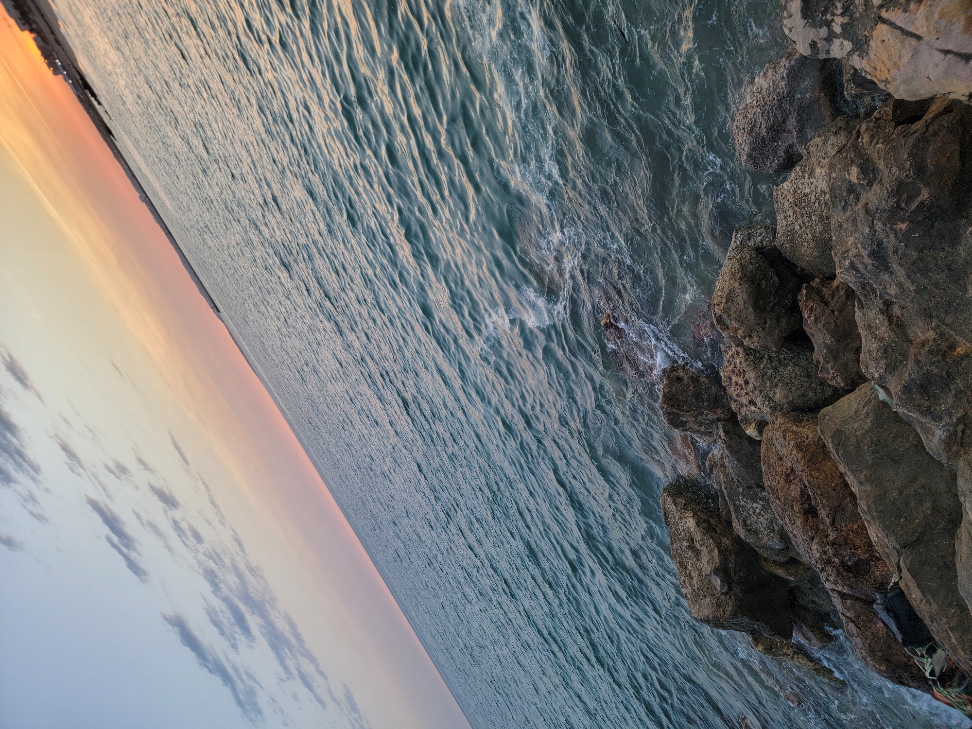 This image captures a coastal scene in Quarteira, Portugal, during a sunset. The ocean occupies the majority of the frame, with gentle waves lapping against a rocky shore in the foreground. The water is a deep teal color, reflecting the twilight sky. The sky itself transitions from a pale blue at the top to a soft pink and orange gradient near the horizon, indicating the setting sun. Wispy clouds are scattered across the upper portion of the sky. The shoreline is lined with large, rough-textured rocks of various sizes, some of which are partially submerged by the water. In the far distance, along the horizon, faint outlines of buildings suggest the presence of the town. There are no people visible in the image, and no specific actions are depicted other than the natural movement of the sea. The overall impression is one of tranquility and natural beauty.