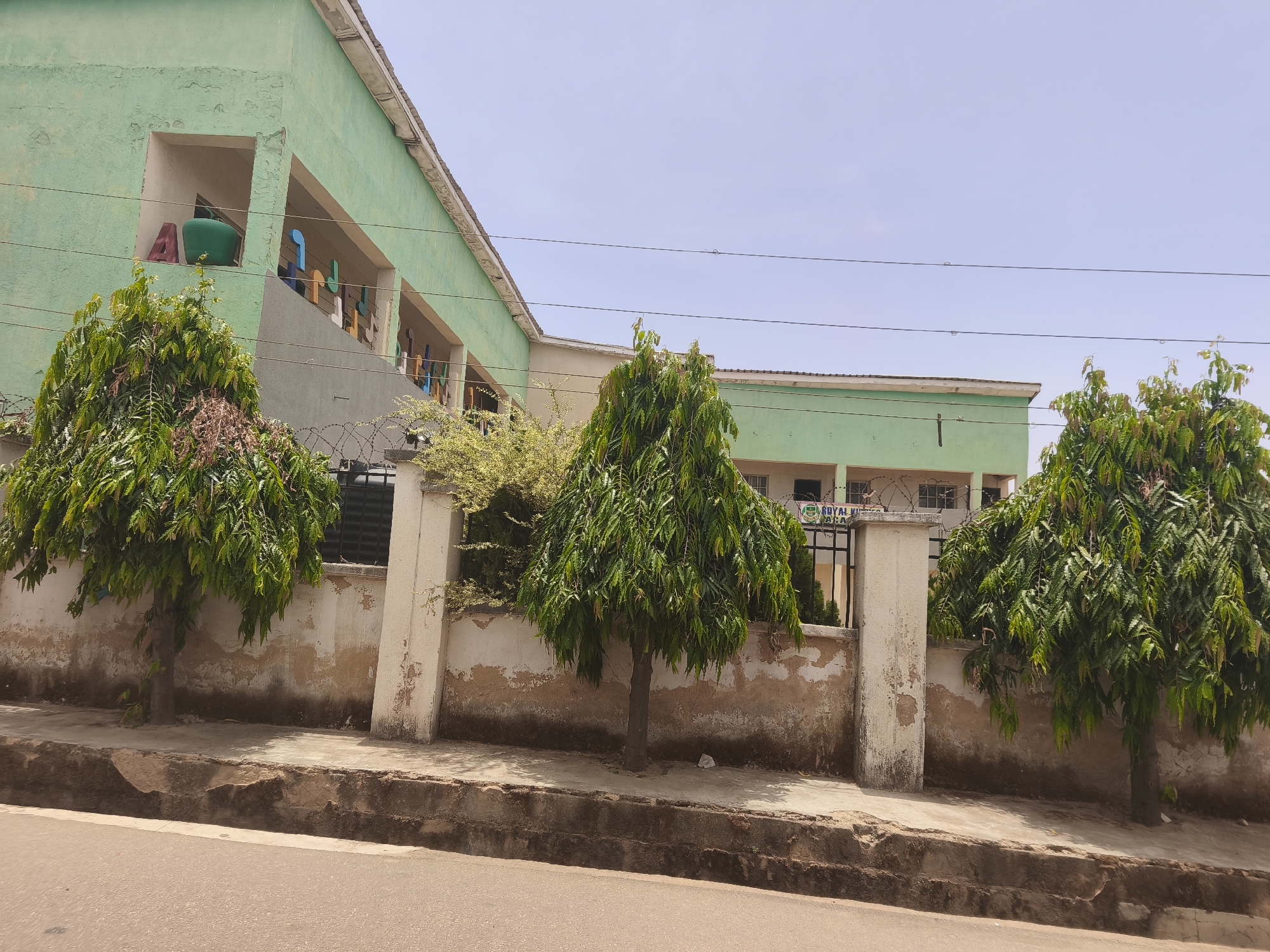 The image depicts a street scene in Kabong, Nigeria, with a light green building in the background. The building's architecture includes recessed areas with colorful objects inside. A weathered wall runs along the edge of the property, punctuated by concrete pillars. Three trees with dense green foliage are planted in front of the wall, partially obscuring it. A cracked and uneven sidewalk is visible, leading to a paved road. Electrical wires stretch across the sky.