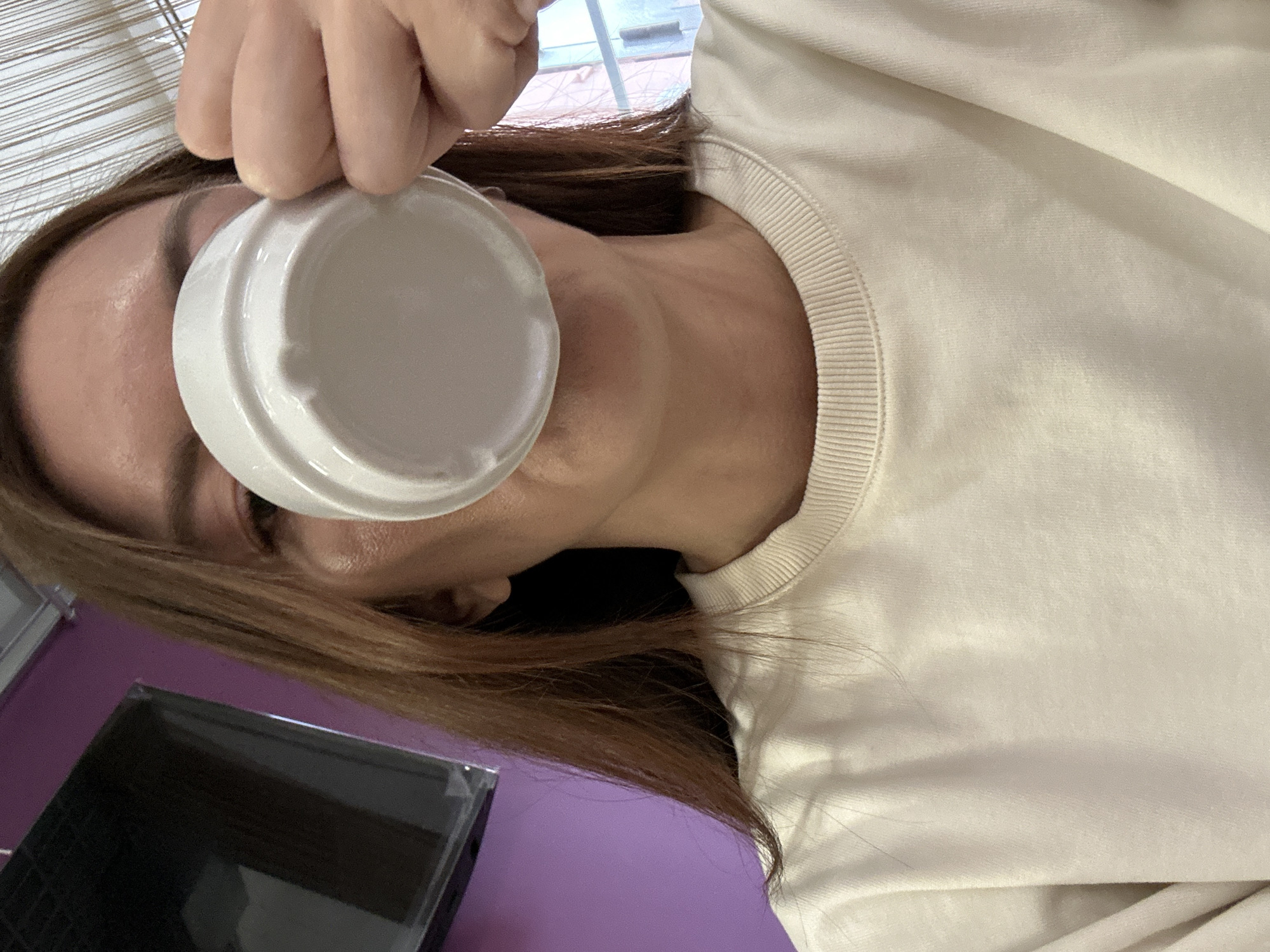A person is holding a white cup in front of their eye. The person has brown hair and is wearing a cream-colored sweater. A black rectangular object is on a purple surface in the lower left corner of the image. The background includes a window with blinds. The location of the image is unknown and cannot be determined from the given image.