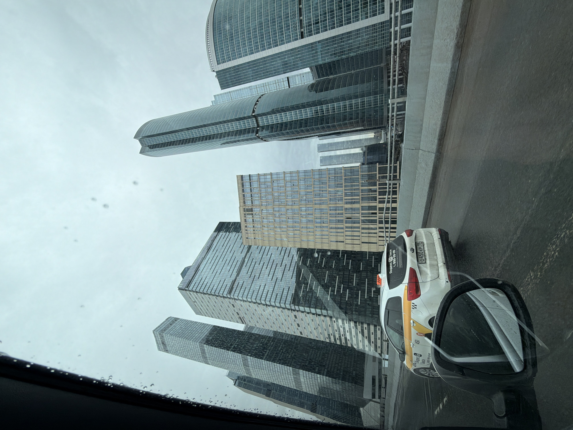 The image is taken from inside a vehicle, looking up at a cluster of tall skyscrapers in Moscow, Russia. The perspective suggests the vehicle is traveling on a raised road or overpass. Rain is visible on the windshield and side mirror. A white taxi is visible to the right of the frame in the adjacent lane. The sky is overcast.