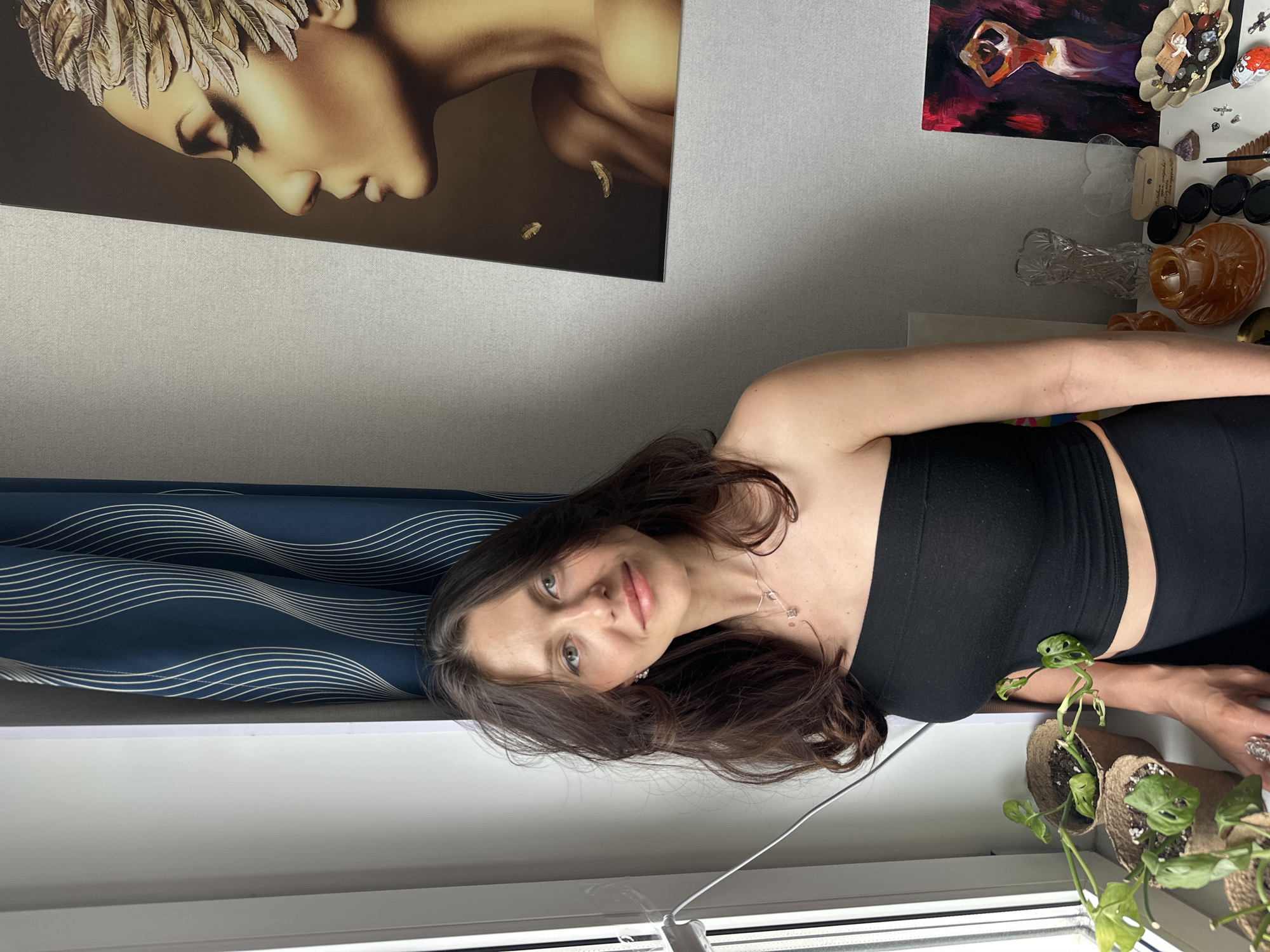 A woman with dark hair and green eyes is lying on a windowsill, wearing a black strapless top. Behind her are dark blue curtains with a white wavy pattern. Above her to the left is a large painting of a woman with feathered headwear. To the right of the painting, and above the woman, is a collage of art, including a painting of a dancing figure and a collection of trinkets and crystals. In front of the woman on the windowsill are several small potted plants. The image was taken indoors in what appears to be a residential setting, possibly in Murino, Russia, based on the request.