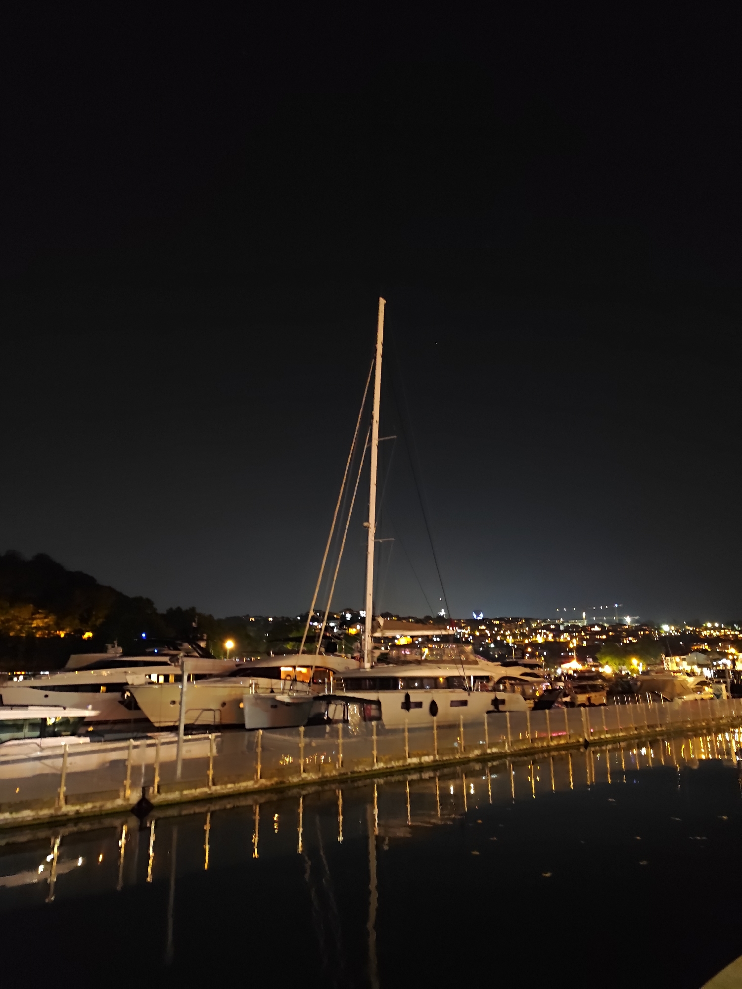 In the city of Sarıyer, Turkey, at night, several docked yachts are illuminated by artificial lights. The yachts are positioned along a waterfront with a railing separating them from the water, which reflects the lights from the vessels and the distant cityscape. The sky is dark, with faint lights visible on the horizon, suggesting a populated area. The scene is one of a tranquil, illuminated harbor at night.