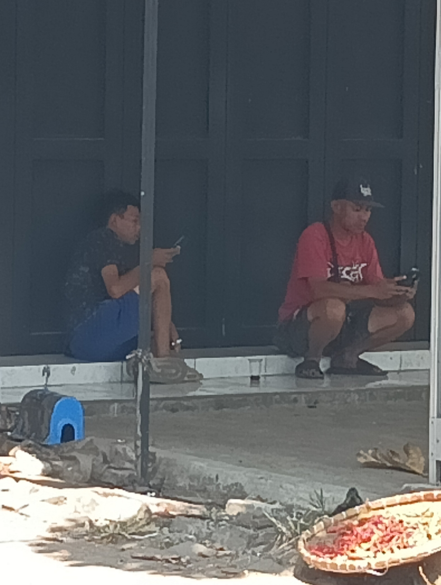 Two men are sitting on the steps outside a building with dark-colored doors. The man on the left is wearing a dark patterned t-shirt and blue shorts, and he is holding a mobile phone in his right hand. He is also holding a cigarette in his left hand. The man on the right is wearing a red t-shirt and dark shorts, and he is wearing a black baseball cap. He is holding a mobile phone in his right hand and appears to be looking at it. A straw basket filled with red items, possibly chilies, is visible on the ground in the foreground. The building appears to be located in Katapang, Indonesia, a busy commercial or residential area.