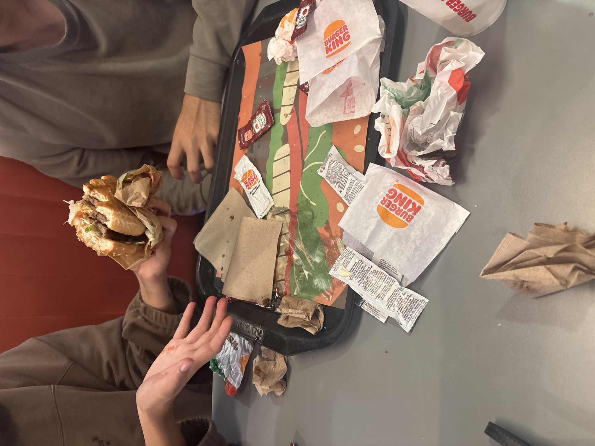 The image depicts a candid overhead view of a fast-food meal at a Burger King restaurant, likely in Puebla, Mexico. Two individuals are partially visible: one wearing a grey hoodie, seen in the upper left with hands near the tray, and another in a brown tracksuit, holding a half-eaten burger with both hands in the lower left.
A black fast-food tray, placed on a grey table surface, occupies the center of the frame. The tray is covered with a patterned paper liner featuring orange, green, and brown stripes. The tray is cluttered with discarded fast-food packaging and remnants of the meal. Numerous crumpled white paper wrappers and bags, all prominently displaying the "BURGER KING" logo in orange, are scattered across the tray. Several condiment packets are also present, including red packets of "TOMATO KETCHUP" and white packets, some also bearing the Burger King branding. Used brown paper napkins and various crumbs contribute to the messy appearance of the tray, suggesting a meal in progress or recently consumed.
In the background, a portion of a red booth or seating area is visible. The lighting is consistent with an indoor restaurant setting. The focus is on the meal itself and the immediate aftermath of dining, with the individuals' faces not shown. The overall impression is a casual, everyday fast-food experience.