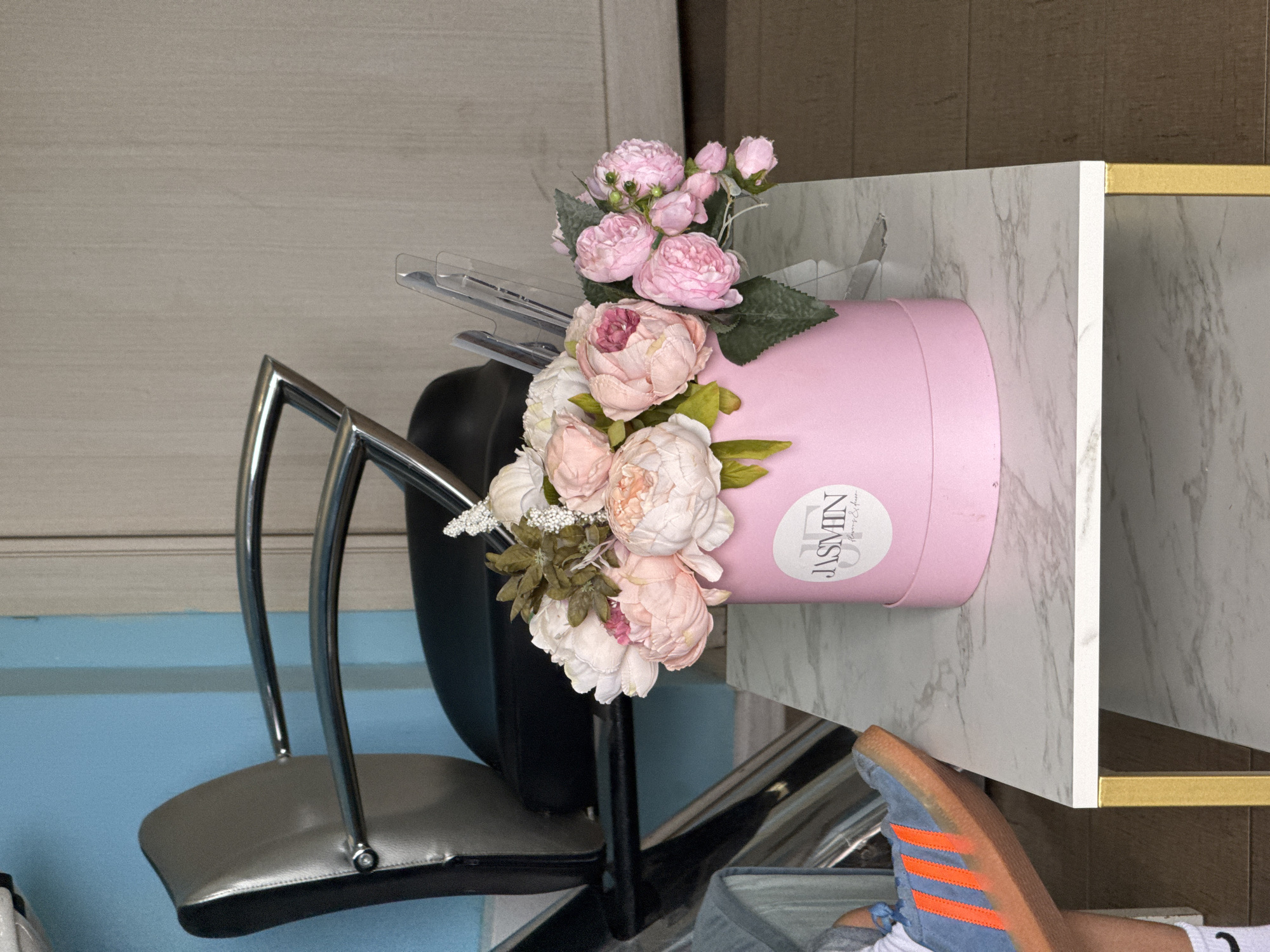In a room with blue and beige walls, a pink floral arrangement sits on a white marble-patterned table. The arrangement features various pink and white peonies and is contained within a pink cylindrical box with a white label that reads "JASMIN Flowers & Decor". Behind the table, a silver and black salon chair with chrome accents is visible. A person's foot wearing a blue sneaker with orange stripes and a white sock is in the lower left corner of the frame, resting on the edge of the table. The scene appears to be indoors, possibly in a salon or a home.