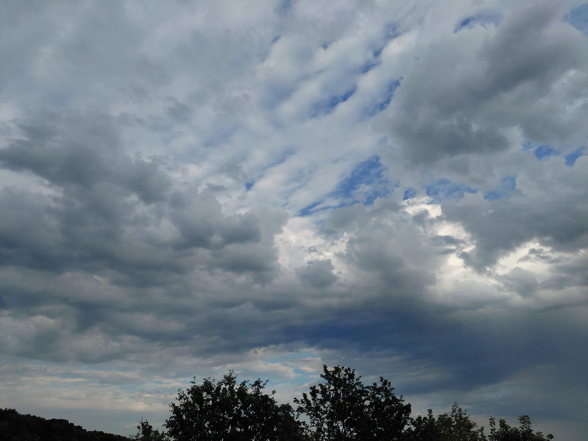 The image depicts a cloudy sky above a tree line, with the horizon visible in the lower part of the frame. The clouds are varied in texture and color, ranging from dark gray to lighter white, with patches of blue sky peeking through in some areas. The trees appear to be silhouetted against the sky, suggesting the image was taken during the day, possibly late afternoon or early evening. There are no people or specific objects identifiable within the scene, beyond the natural elements of the sky and vegetation. The overall atmosphere is overcast and somewhat dramatic due to the cloud formations. The location is stated to be Verhnyaya Podstyopnovka, Russia.