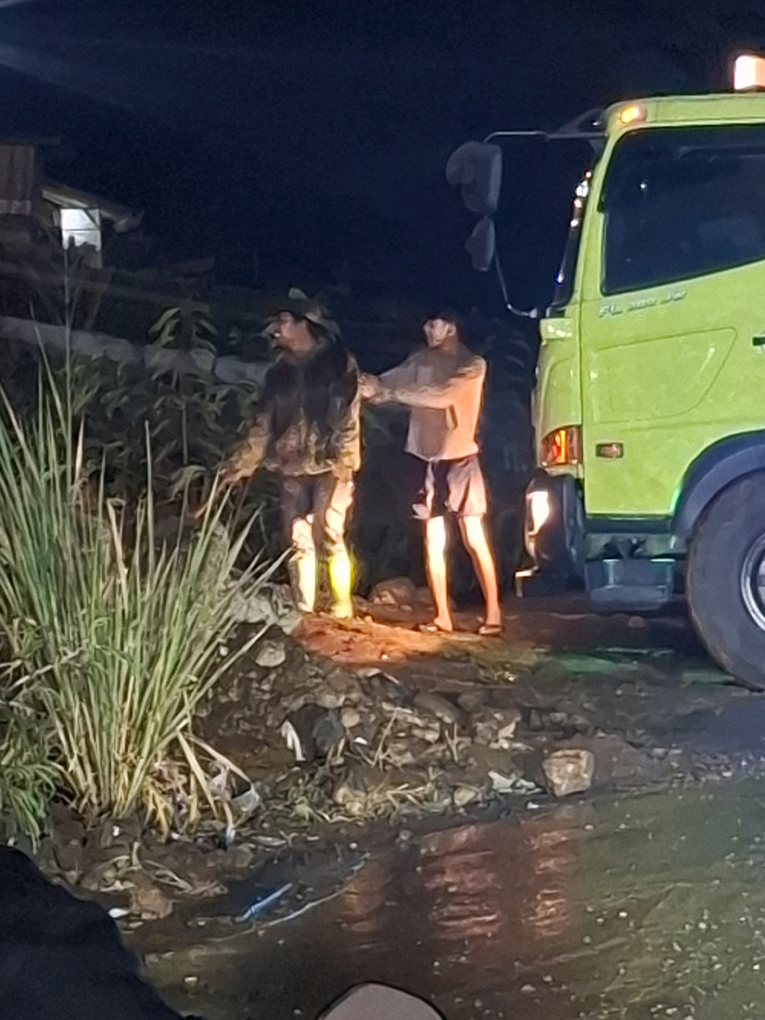 In Ciheulang Tonggoh, Indonesia, at night, two men are near a bright green truck. One man, wearing a camouflage jacket and hat, stands by a pile of rocks and dry grass, his right hand extended towards the truck. The other man, in a light-colored hoodie and dark shorts, stands closer to the truck, his arms outstretched. The ground is uneven, with water and debris visible. The truck has flashing orange lights on its roof and appears to be a large vehicle. The background shows a dimly lit building and vegetation.