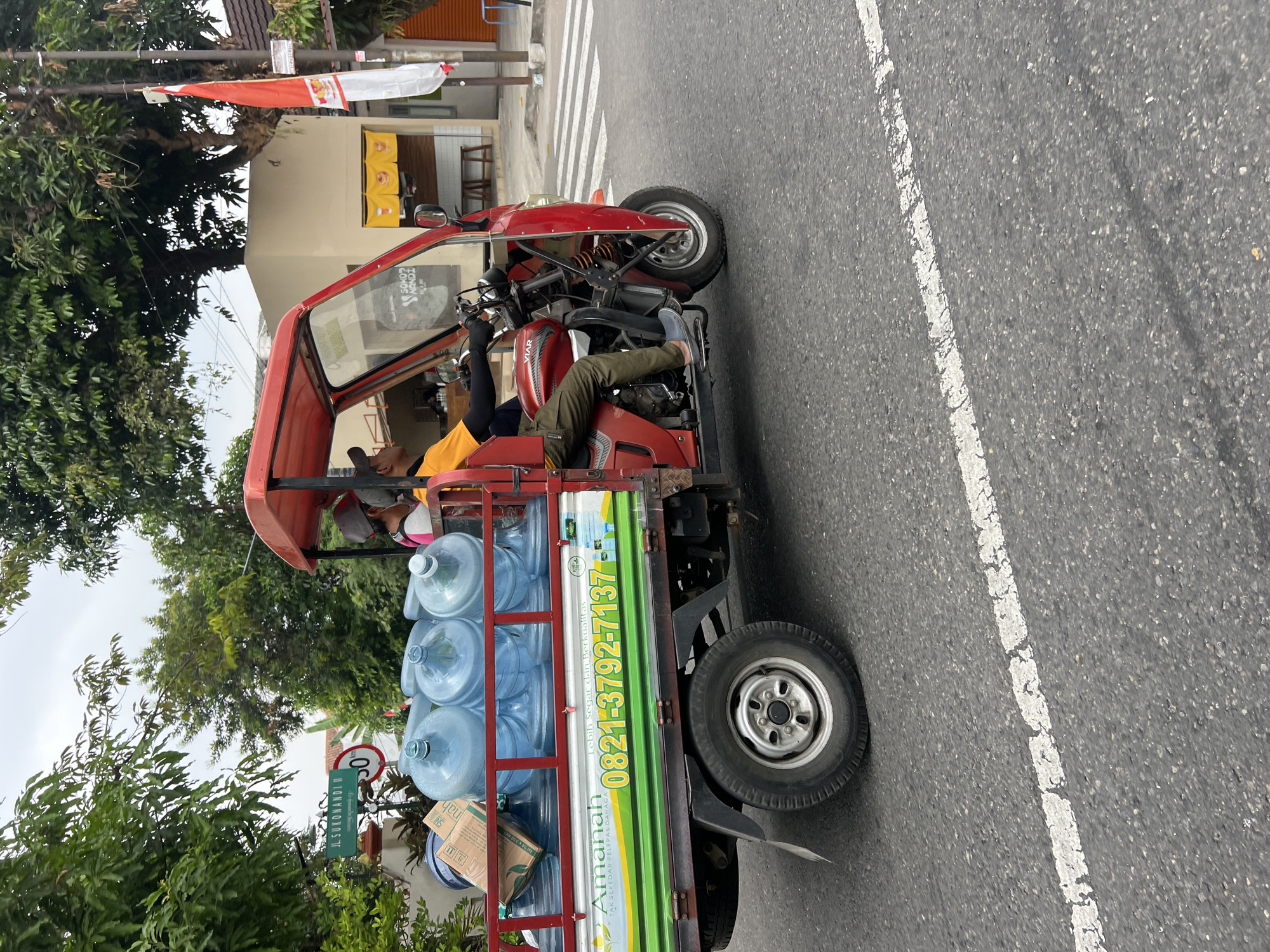 A red, three-wheeled vehicle is driving on a paved road in Yogyakarta, Indonesia. The vehicle is carrying several large, clear water jugs in its rear cargo area, along with some cardboard boxes. Two men are seated in the vehicle; one is driving, and the other appears to be a passenger. The driver is wearing a yellow shirt and green pants, while the passenger is wearing a grey cap and a pink shirt. A red canopy covers the passenger compartment. In the background, there are trees, buildings, a street sign that reads "Jl. Sukonandi," and a red and white Indonesian flag. The road has white lane markings.