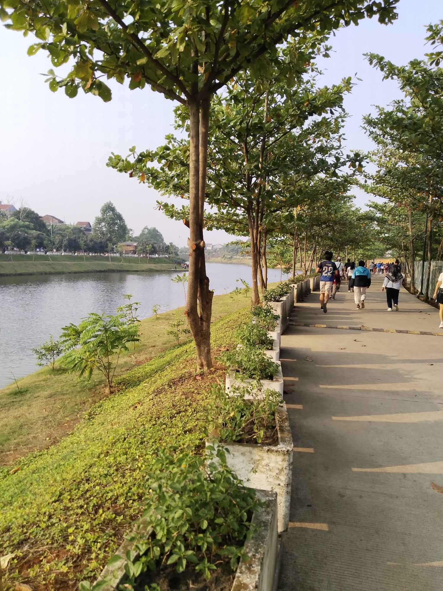 This image depicts a paved walkway alongside a body of water, likely a river or canal, in South Jakarta, Indonesia. The walkway is lined with trees casting shadows on the pavement. Several individuals are walking or jogging on the path, suggesting a public recreational area. The environment appears to be a park or a developed waterfront area, with green grass and vegetation along the embankment. In the background, buildings and more trees are visible. The overall scene suggests a pleasant outdoor environment for exercise and leisure in an urban setting.