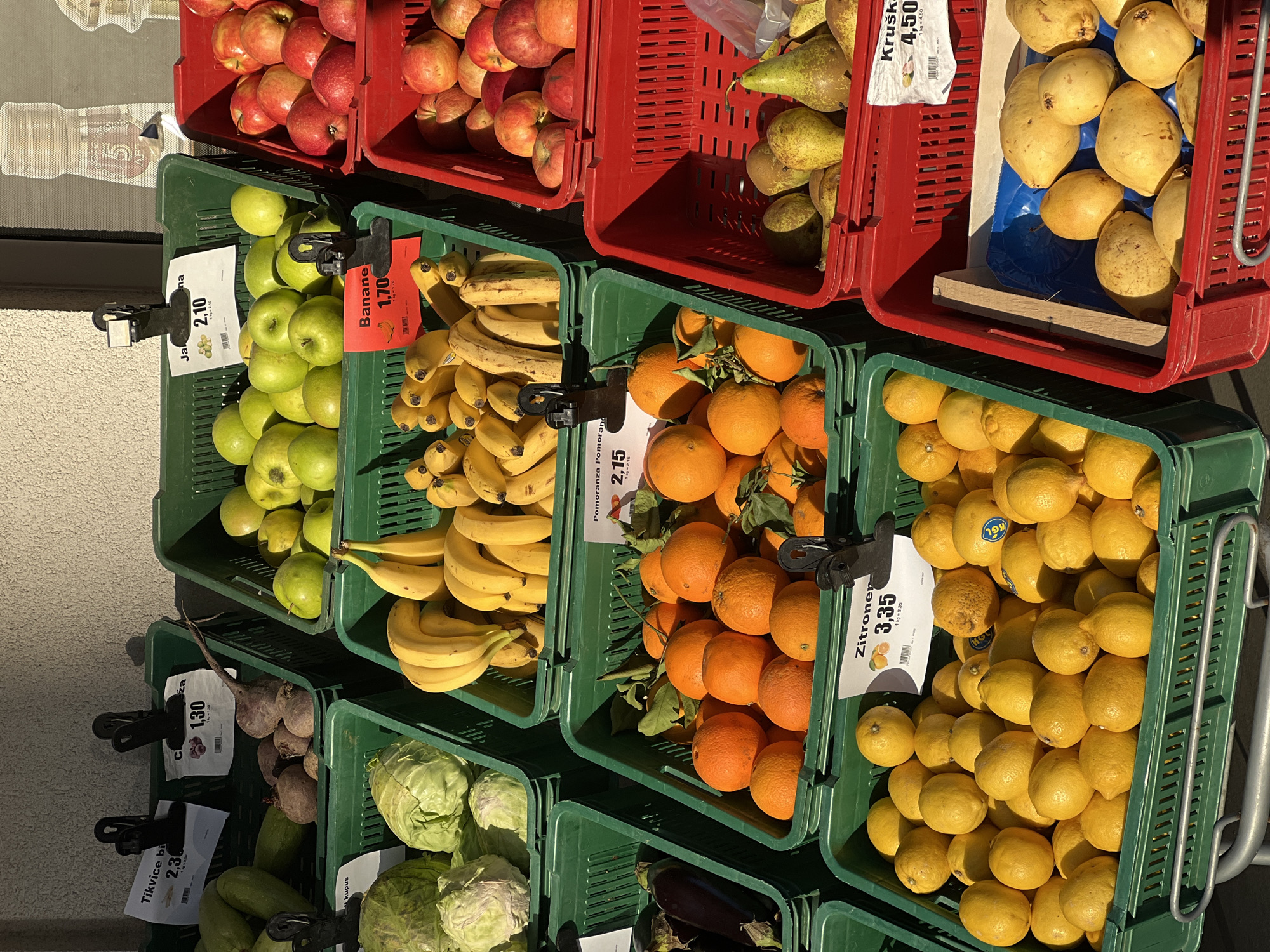 The image shows fruit and vegetables for sale, displayed in red and green plastic crates. The crates are arranged in stacks. Top left are red apples, with green apples below them. To the right of the apples are bananas. There are oranges to the right of the bananas, and lemons on the right of the oranges. Above the bananas, oranges and lemons are pears and more red apples. Under the green apples, bananas, oranges and lemons are more produce including beets, cabbage, zucchini and eggplant. Price tags in German are visible on some of the produce. The location may be Rafailovići, Montenegro.
