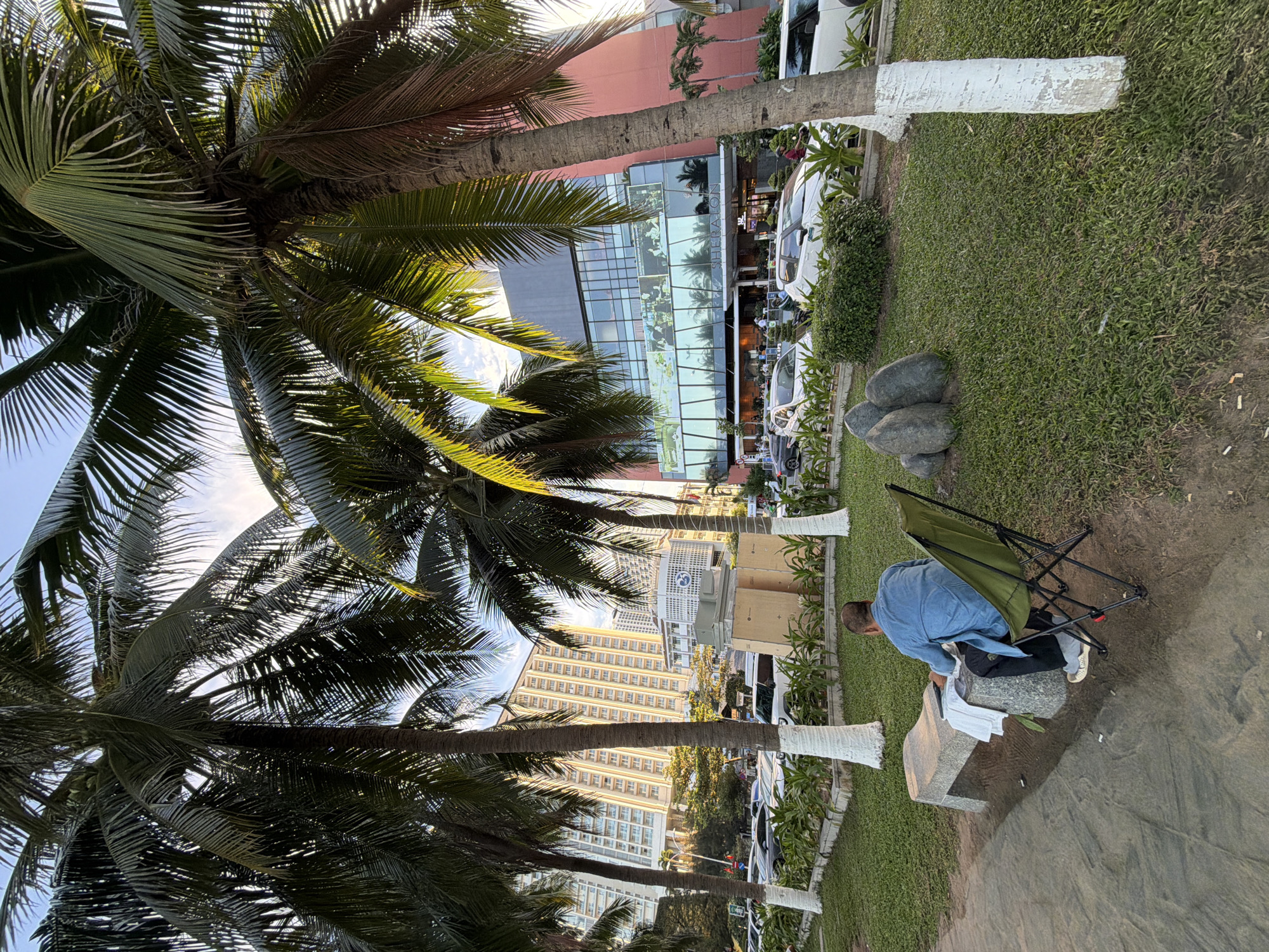 The image shows a man sitting on a green camping chair in front of a stone bench, facing a large building with a glass facade. He is wearing a blue denim shirt and black pants, and appears to be reading or working on a laptop. In the background, palm trees line a grassy area with decorative stones and bushes. Beyond the trees, a modern hotel with a red and glass exterior is visible, along with a street filled with cars and motorbikes. The scene is likely in Nha Trang, Vietnam, given the presence of palm trees, coastal architecture, and the busy urban environment. The man is positioned on a patch of sandy ground next to a well-maintained grassy area.
