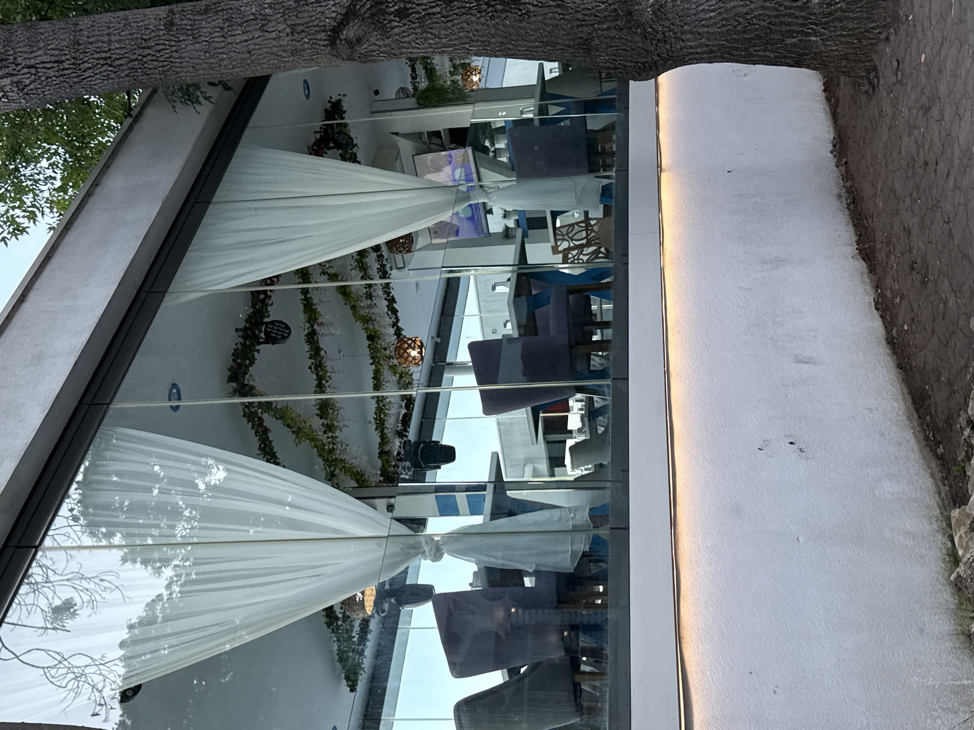 The image depicts the exterior of a modern establishment, likely a restaurant or cafe, with large glass windows. Through the windows, several tables and chairs are visible, suggesting an indoor seating area. White sheer curtains hang inside, partially obscuring the view. Decorative green garlands are draped across the ceiling. Multiple pendant lights with a warm glow are also visible. A large tree trunk is prominent on the right side of the frame, obscuring part of the building's facade. The ground in front of the establishment appears to be a mix of dirt and possibly pavement. A white wall runs along the bottom of the image, illuminated by a strip of light. The overall impression is one of an urban setting with an attempt at a contemporary and inviting ambiance.