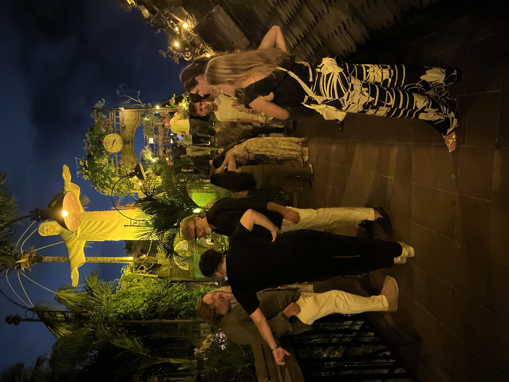 This image depicts an outdoor scene at night, likely in Kuta, Indonesia, featuring a gathering of people on a walkway. Dominating the background is a large, illuminated statue resembling Christ the Redeemer, surrounded by lush greenery and palm trees, suggesting a tropical setting. In the middle ground, an arched structure with a prominent clock is visible, adorned with flowering vines. Several individuals are interacting in small groups along the walkway, which is paved with dark tiles. The lighting is dim, with artificial lights illuminating the statue, foliage, and some areas of the walkway, creating a warm and inviting ambiance. The overall atmosphere appears social and relaxed.
